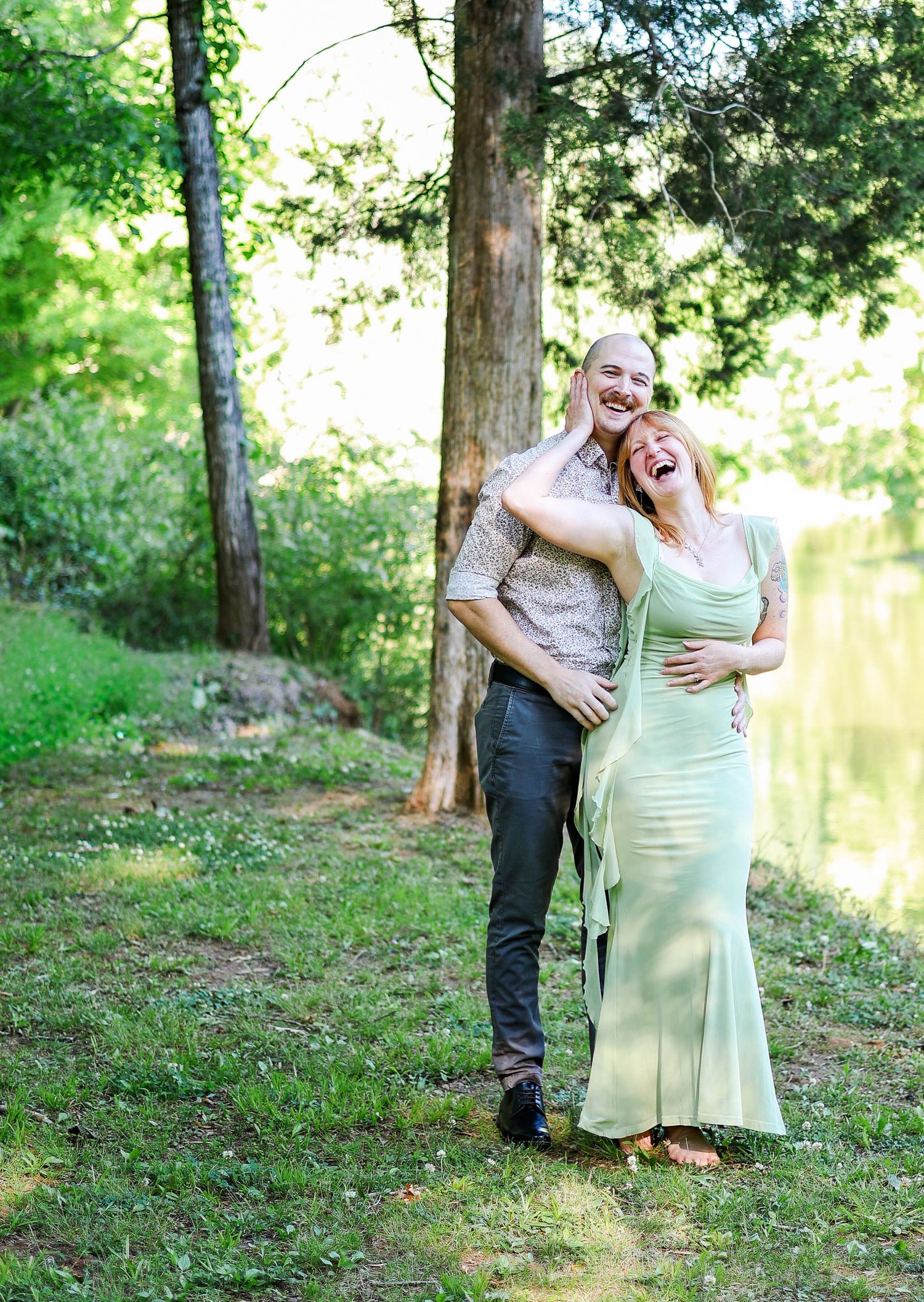 Laughing couple embracing outdoors in lush green park setting, woman in mint dress, man in floral shirt.