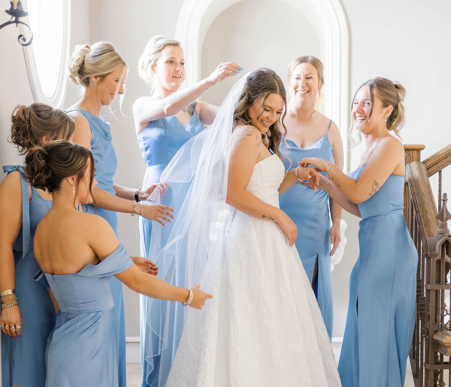 Bride in white gown surrounded by smiling bridesmaids in blue dresses adjusting her veil on a staircase.