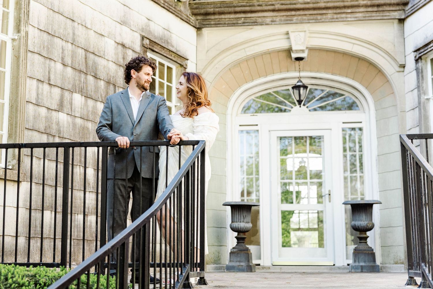 Elegant couple in formal attire standing on stone steps outside a classic building with arched windows.