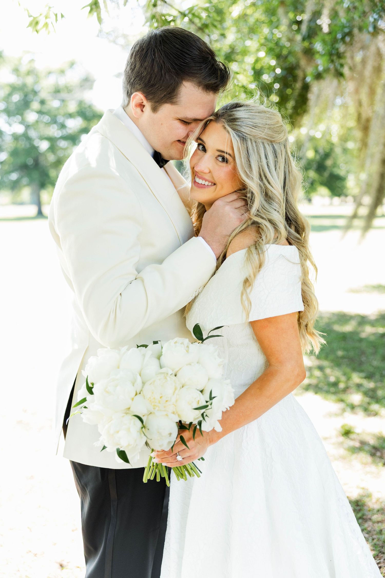 A bride holding white peonies embraces her groom in a white tuxedo jacket outdoors under lush green trees.