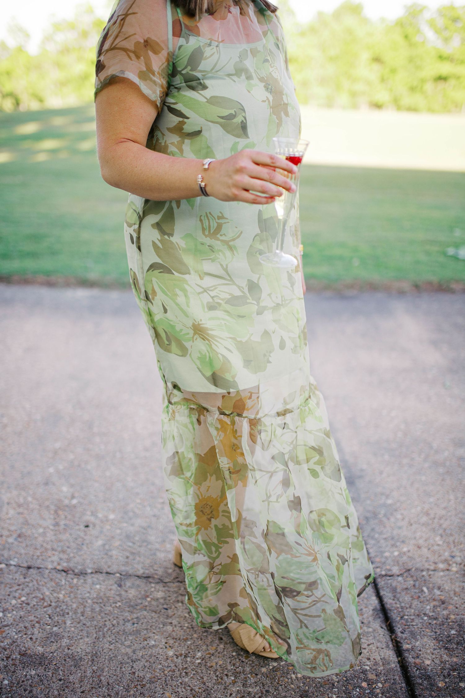 Woman in a flowing green floral maxi dress holding a glass of red wine outdoors at a garden event.