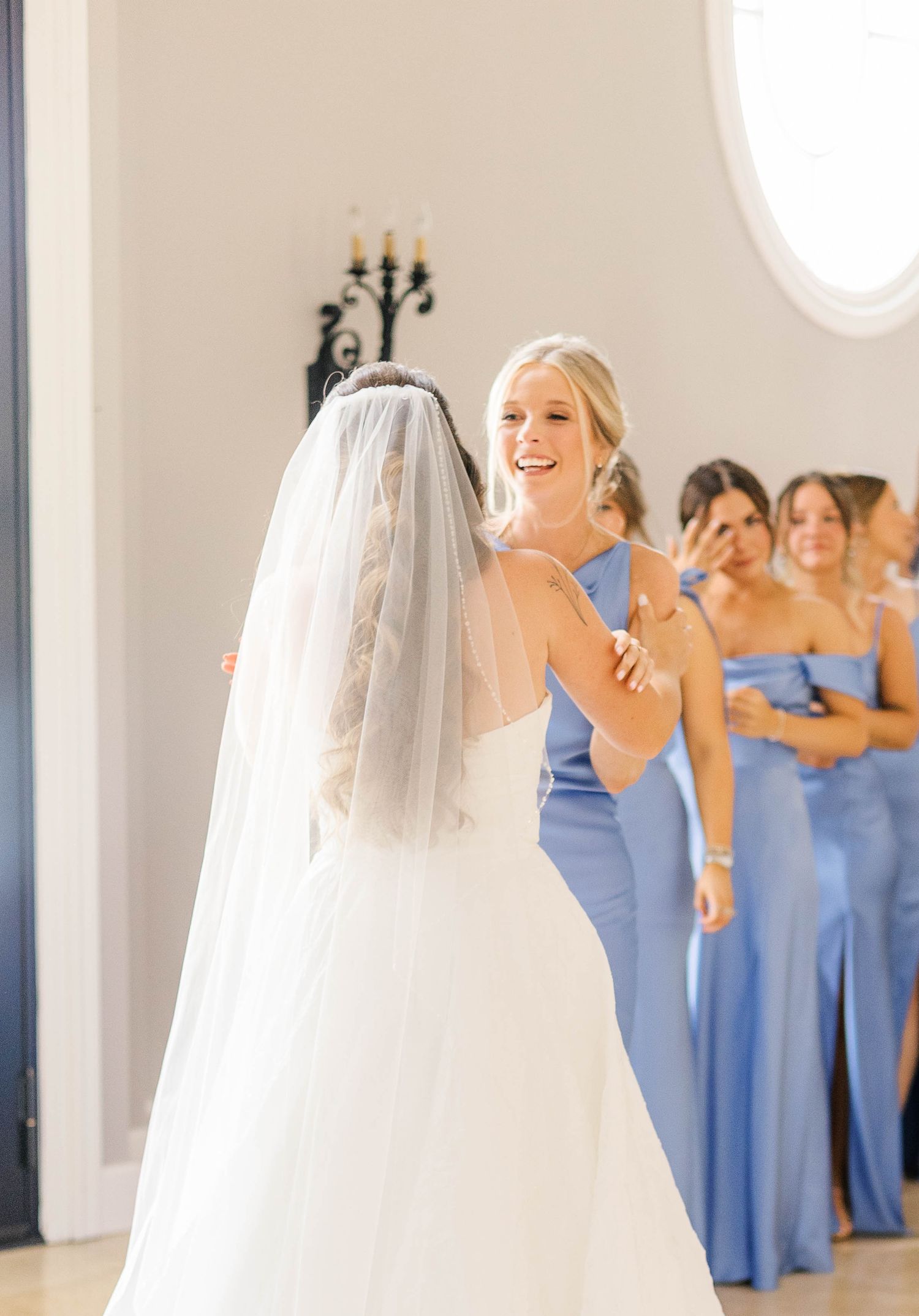 Bride in white gown and veil shares a joyful moment with bridesmaid in blue dress, with bridal party in background.