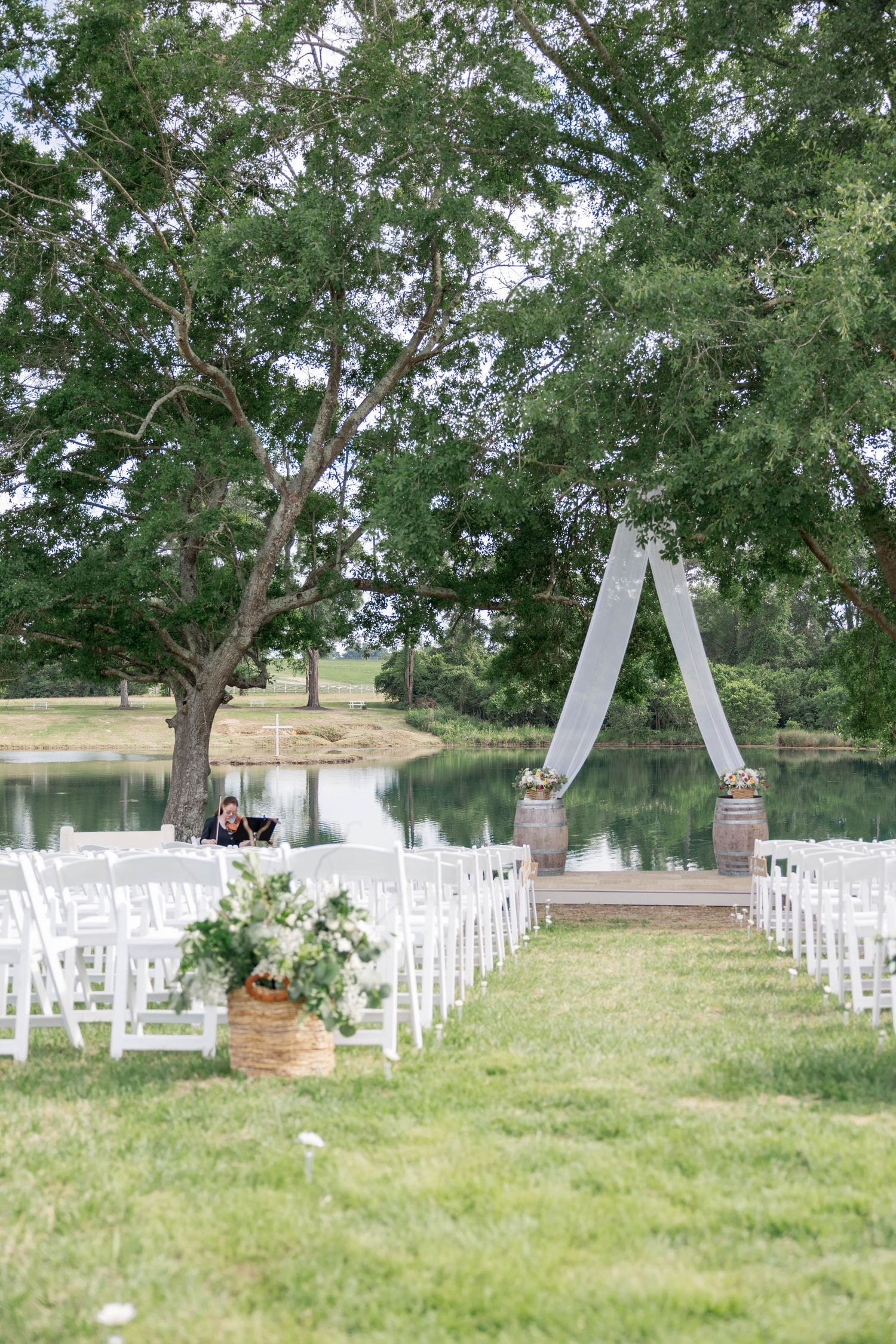 Outdoor wedding ceremony setup with white chairs, floral arch, and a serene pond backdrop surrounded by lush green trees.