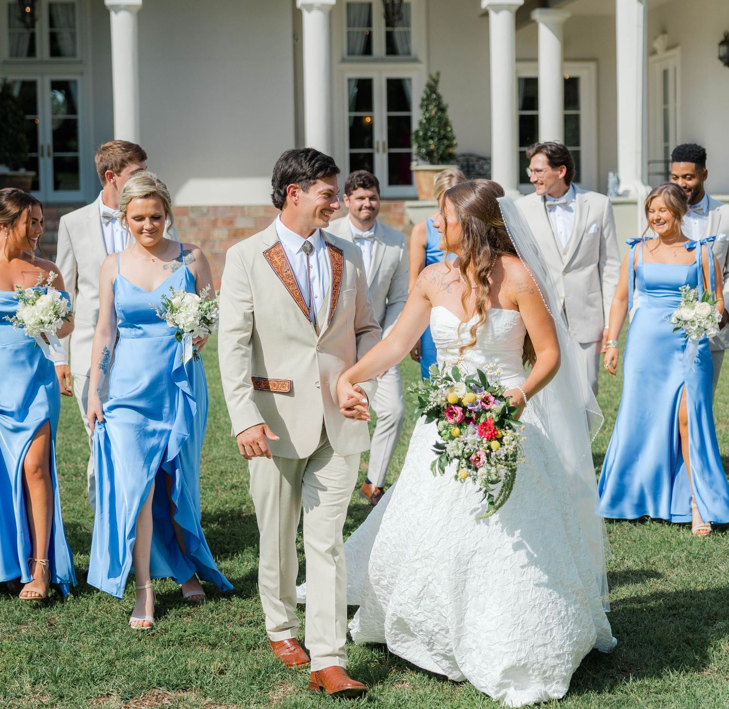 Bride and groom hold hands smiling, surrounded by wedding party in blue gowns and tan suits on sunny lawn.
