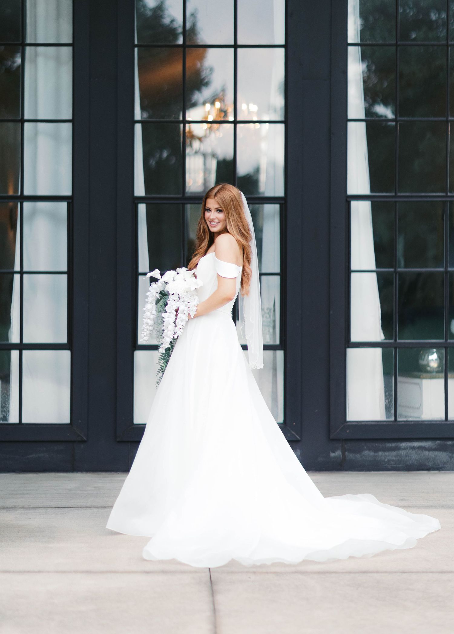 Redhead bride in off-shoulder white ball gown with long train poses before black glass doors.