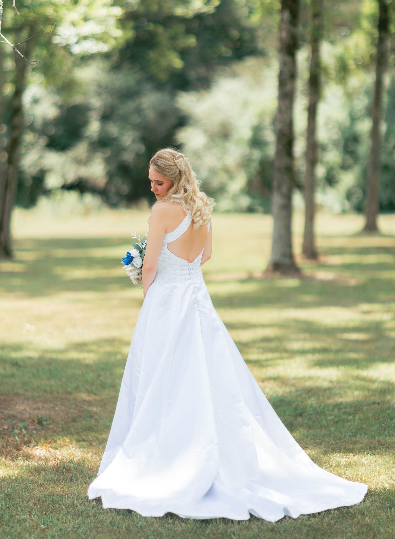 Bride in white backless wedding gown with train holds bouquet outdoors among green trees.