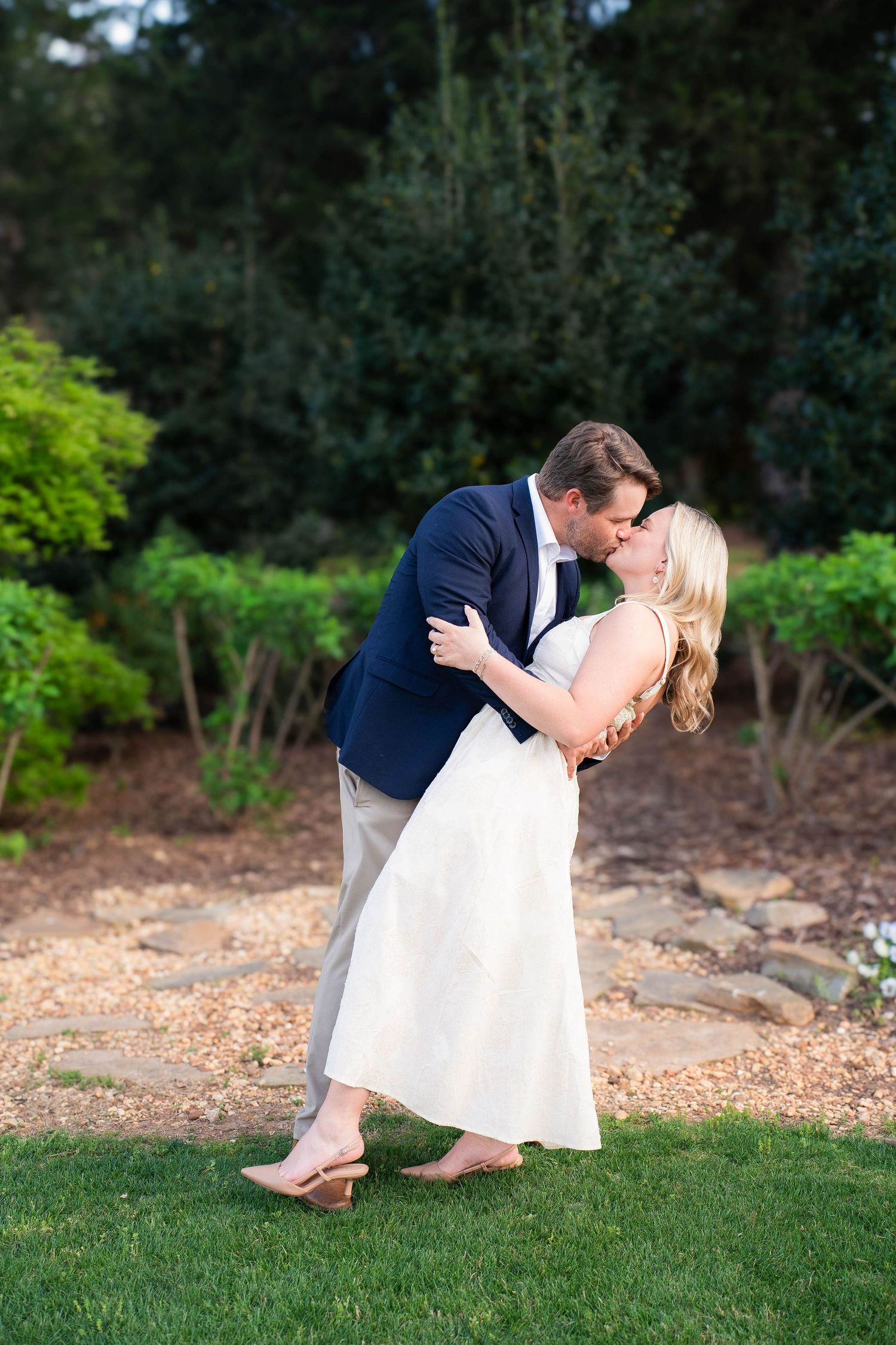 A groom dips his bride for a romantic kiss in a lush garden setting, she wearing a white dress and nude heels.