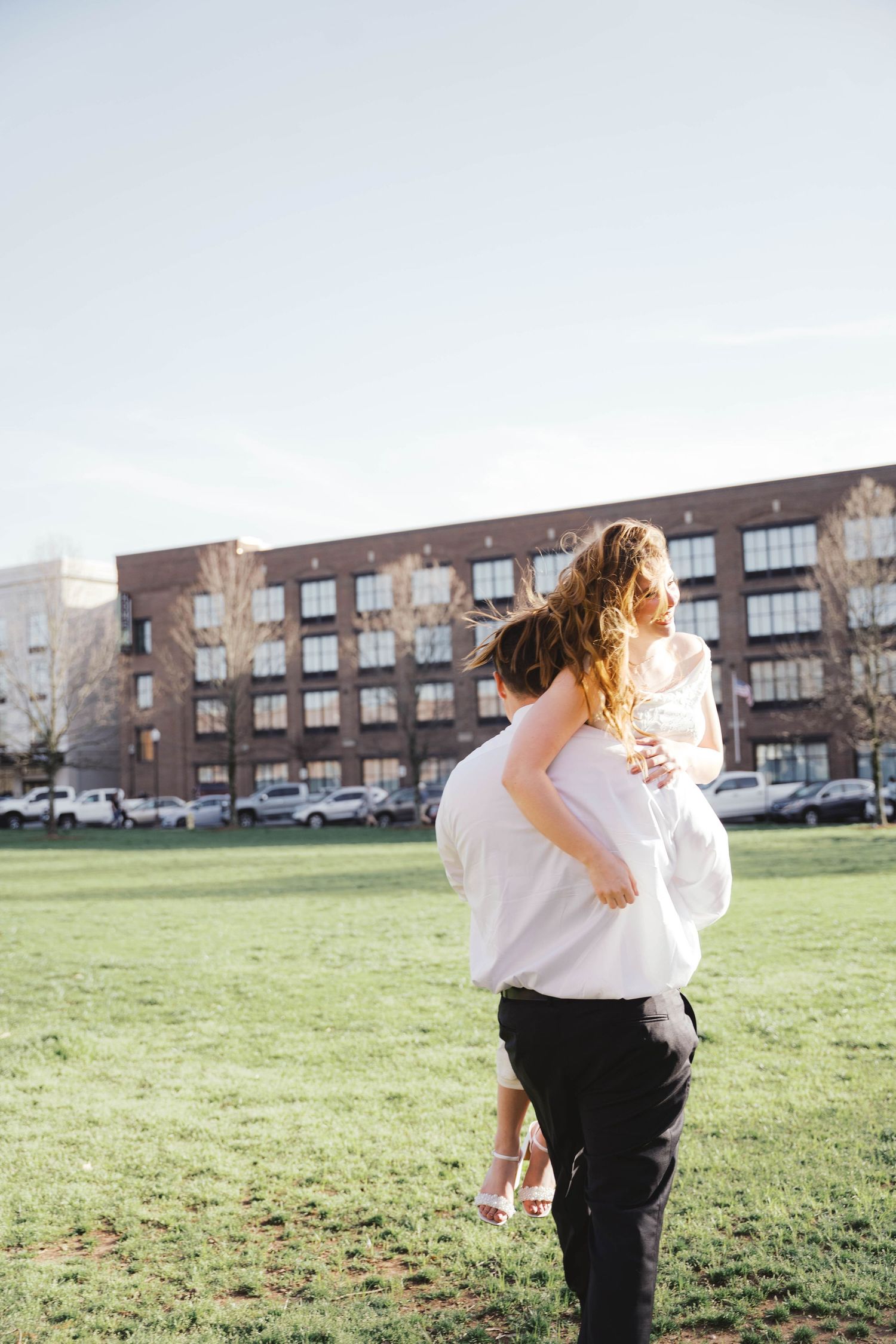 A man carries a woman on his back across a green lawn near a brick building on a sunny day.