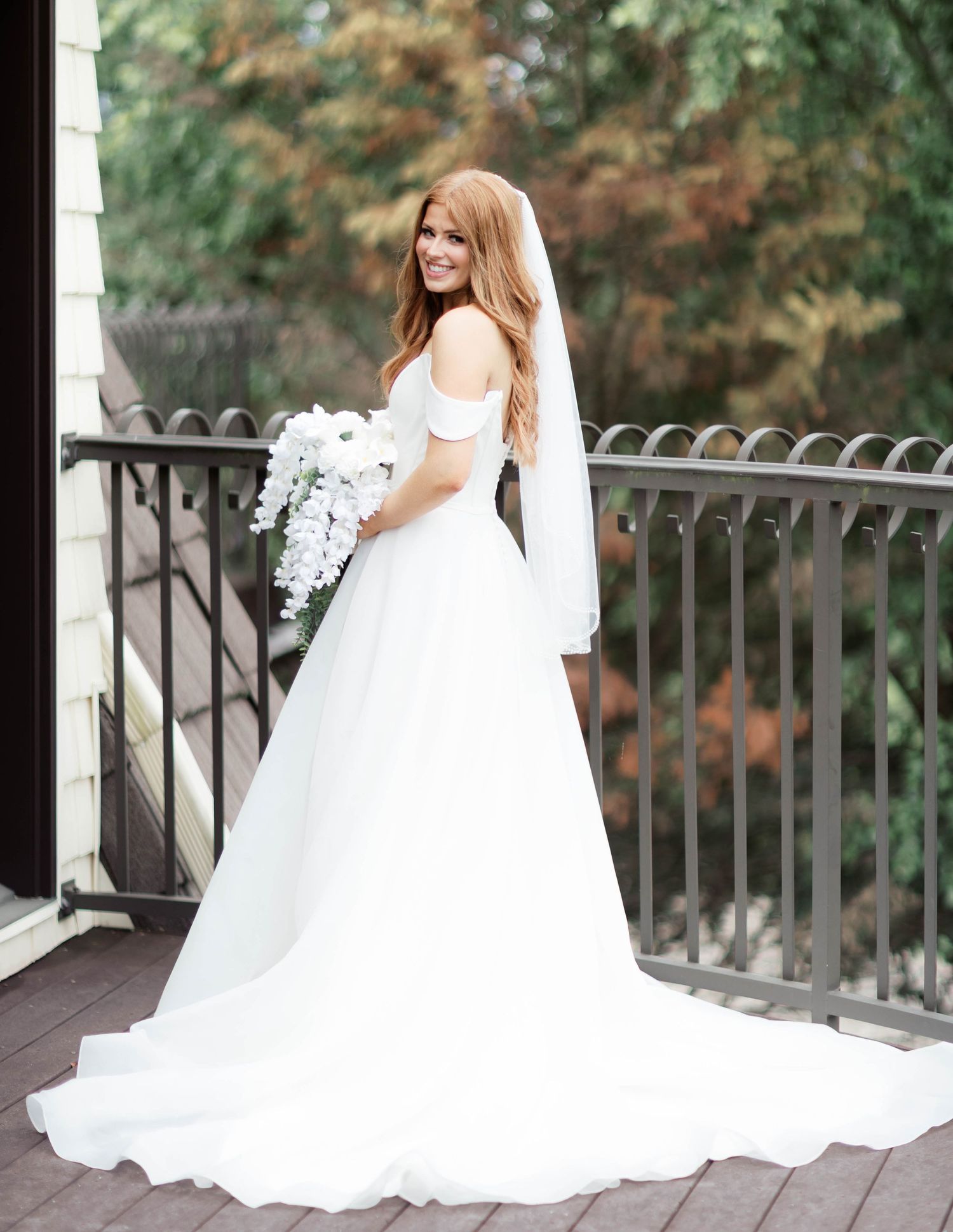Bride in off-shoulder white gown with veil holds white bouquet on balcony, smiling over shoulder.