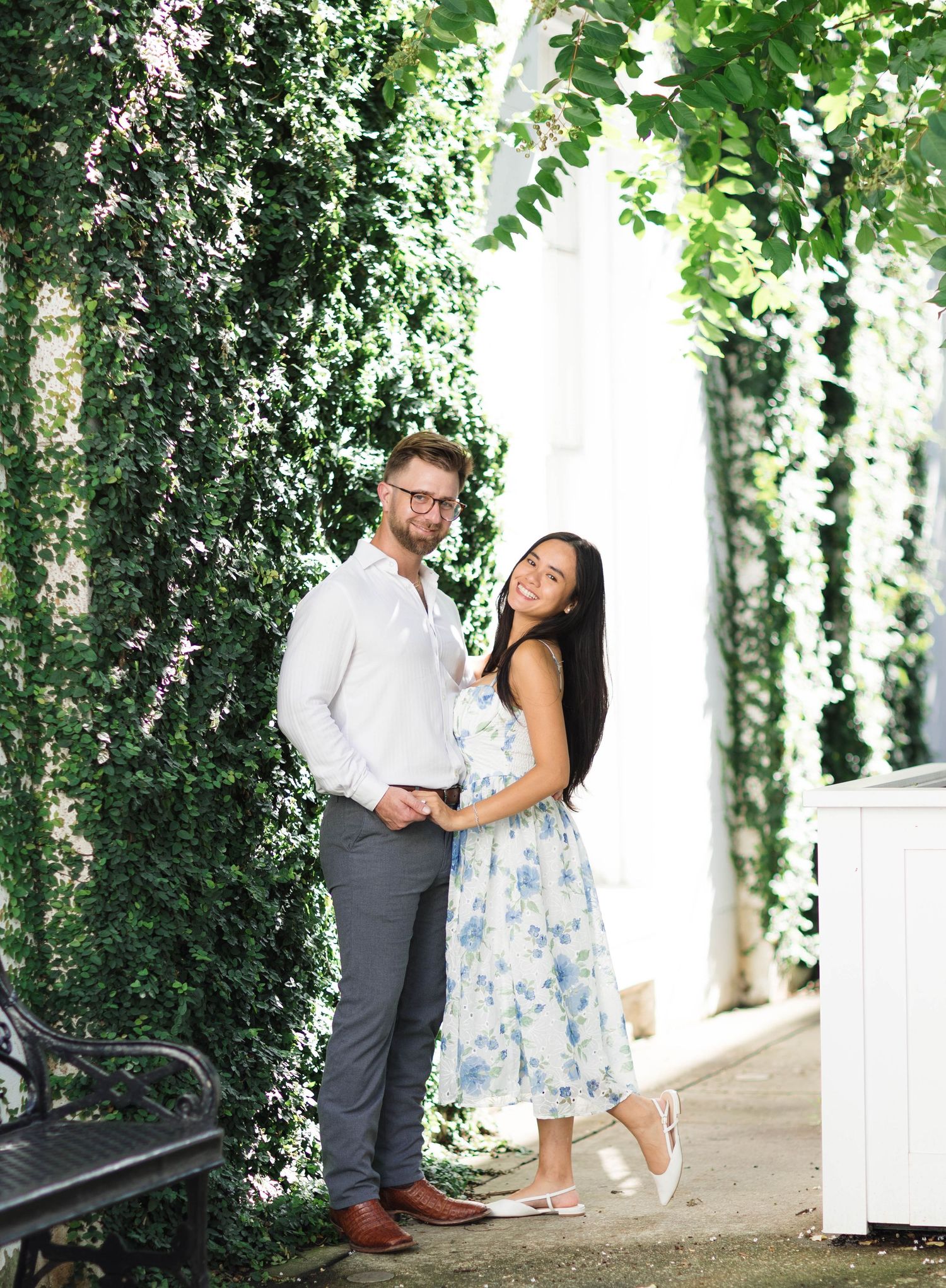 Couple posing together in front of lush ivy-covered wall, woman in floral dress, man in white shirt and gray pants.