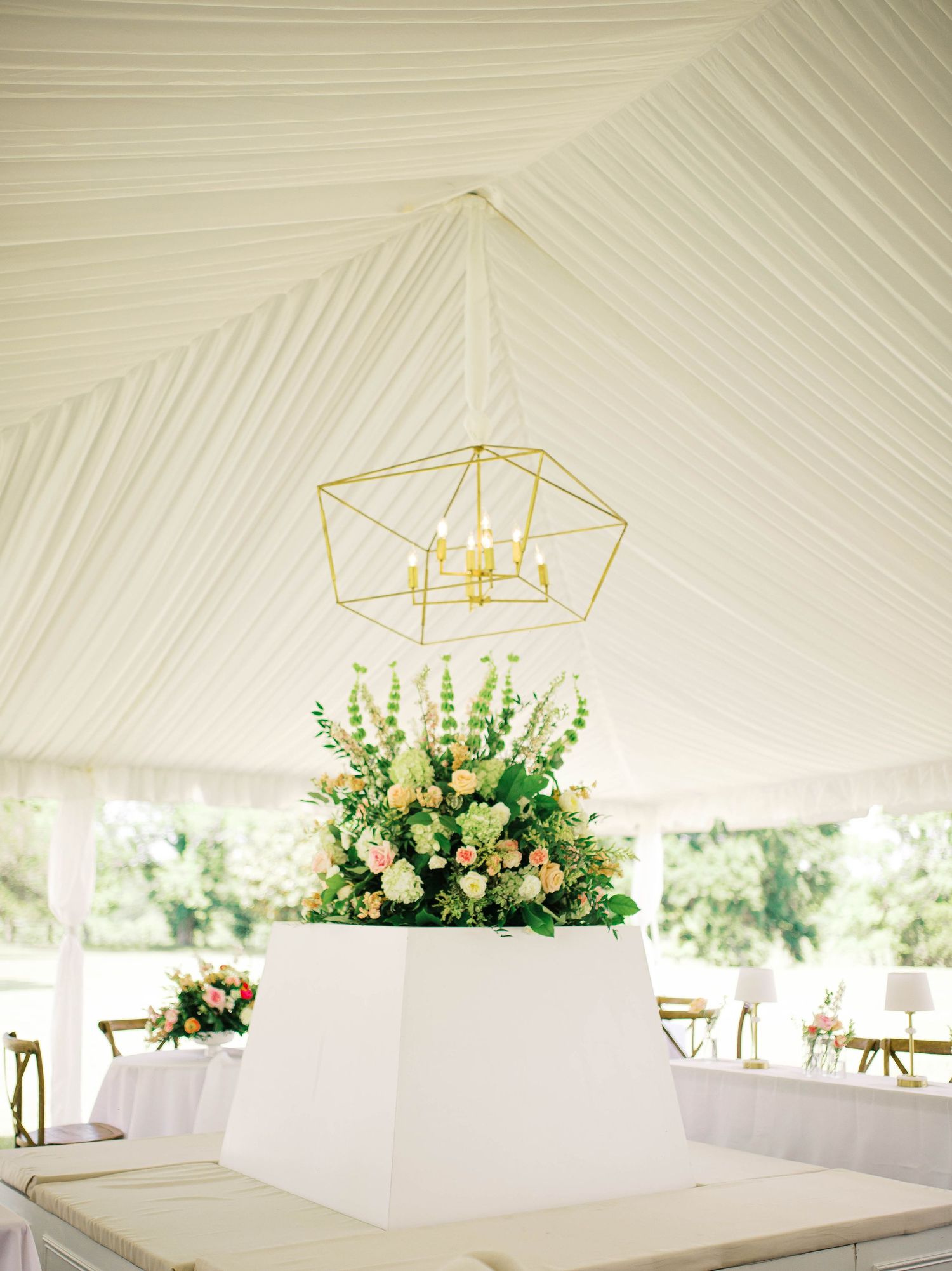 Gold geometric chandelier hangs above a lush floral arrangement on a white pedestal inside an elegant white draped tent.