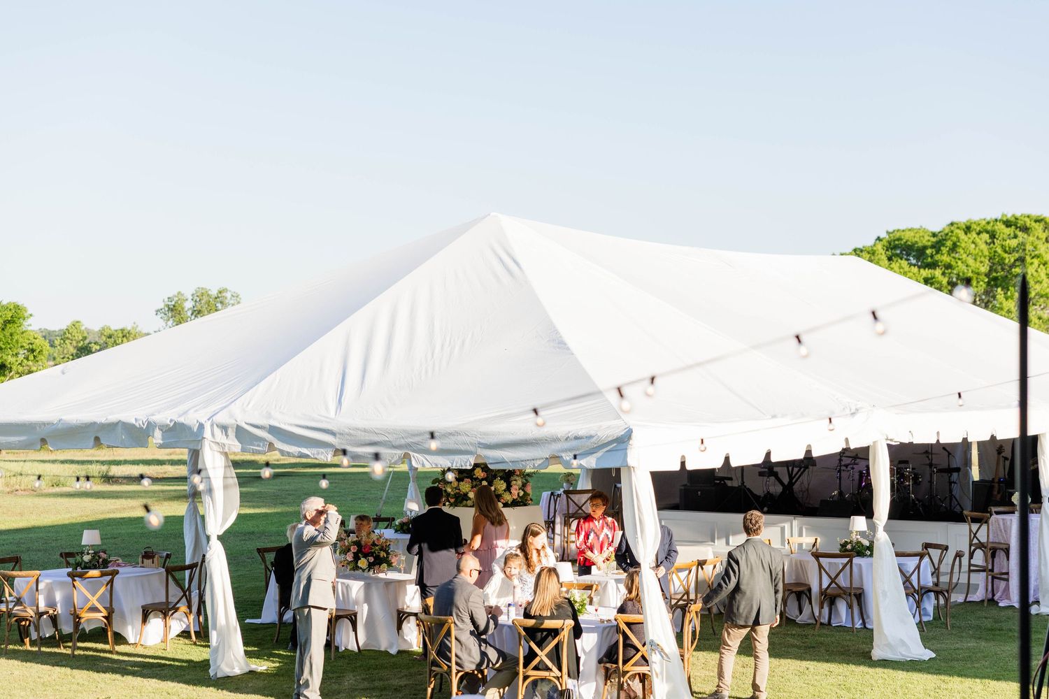 Elegant outdoor wedding reception under a large white tent on a green lawn with string lights and floral centerpieces.