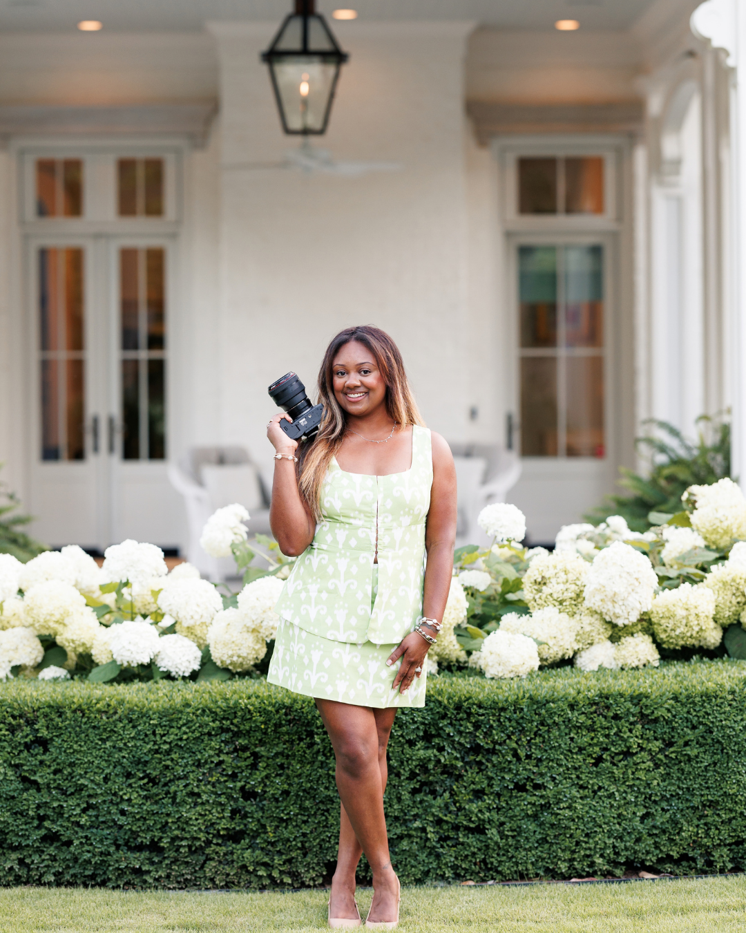 A smiling woman in a green floral dress holds a camera in front of white hydrangeas at an elegant estate.