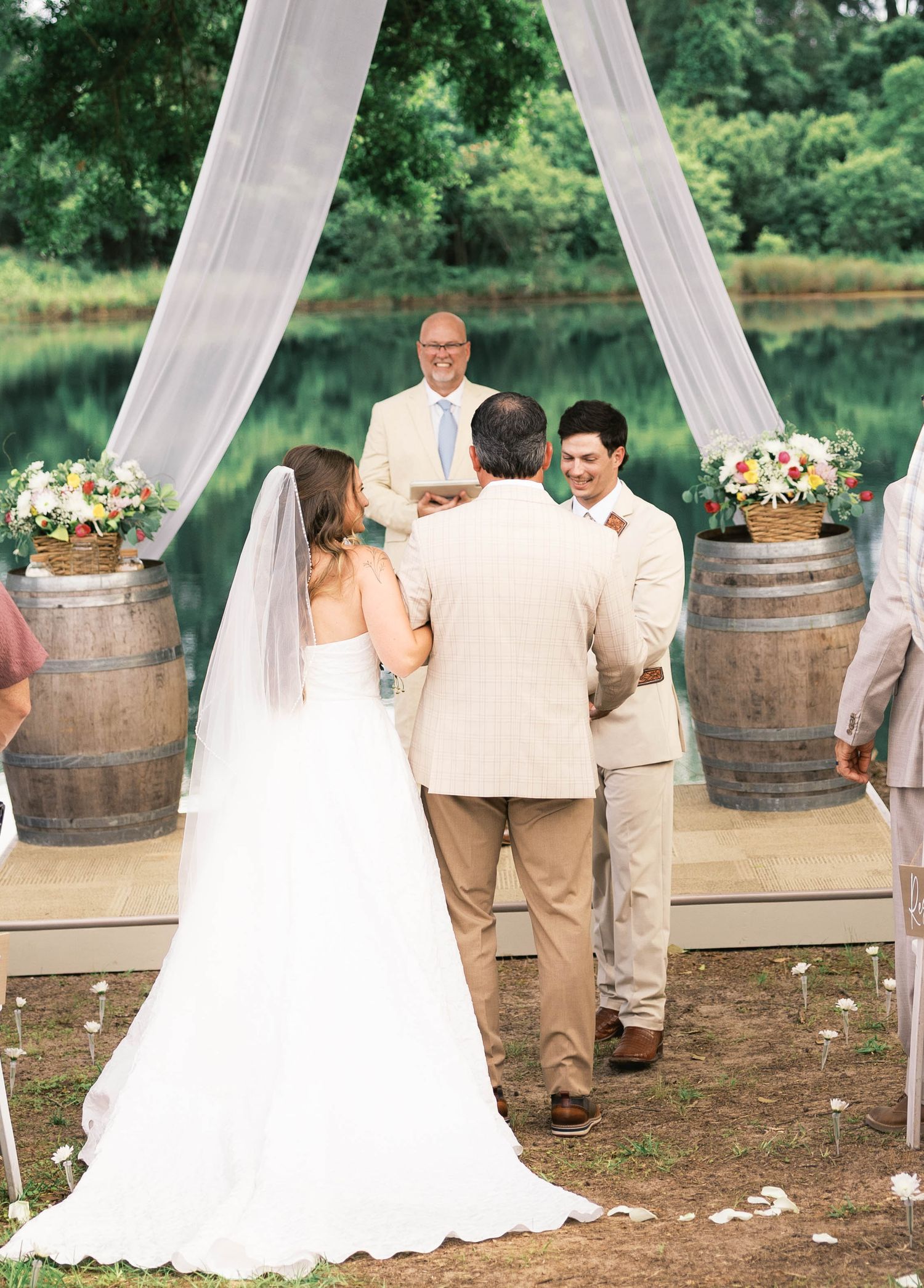 Outdoor wedding ceremony with bride and groom at rustic barrel arch draped in white fabric near a serene pond.