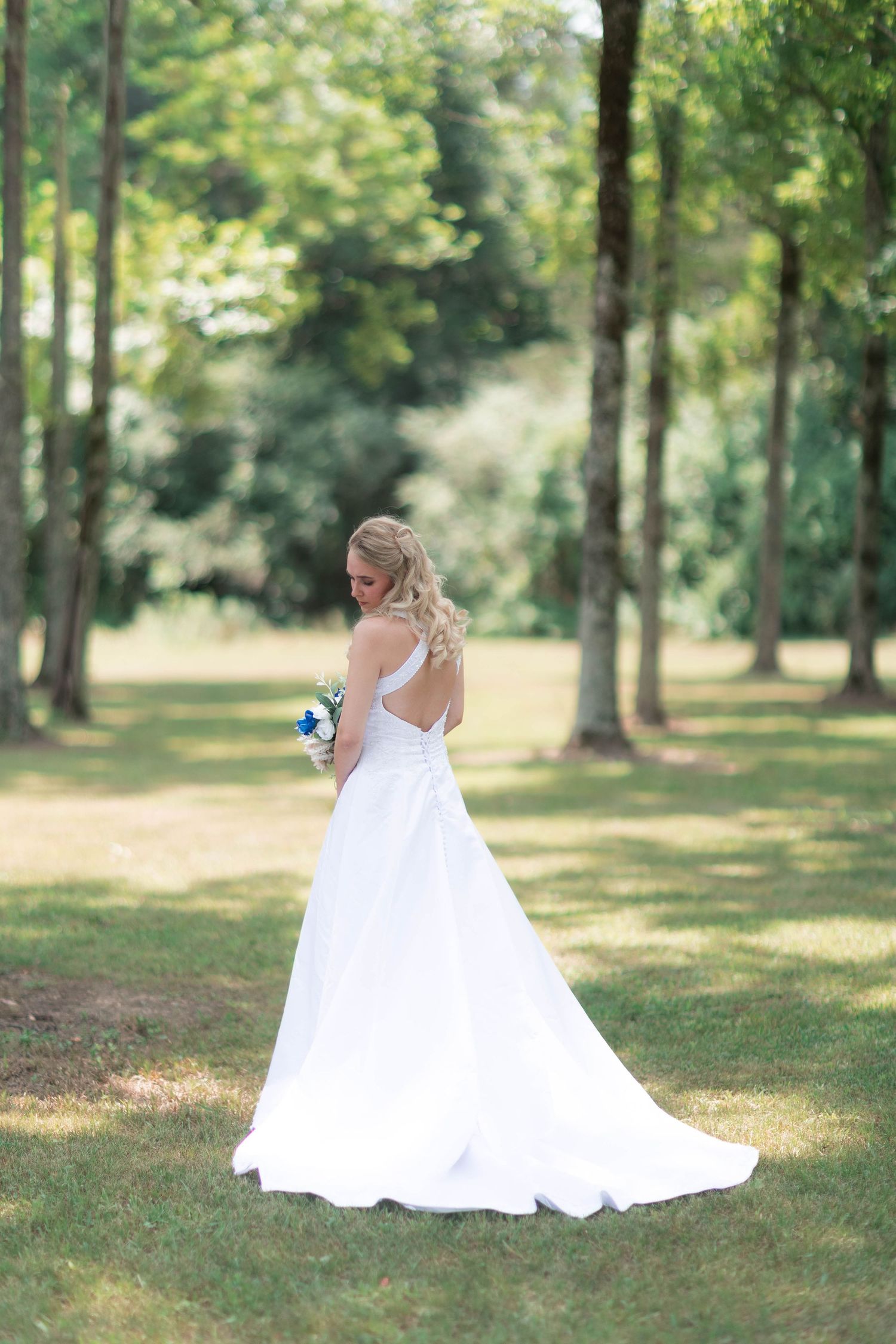 Bride in white gown with train holds bouquet, standing in sunlit green park, showing elegant open back dress detail.