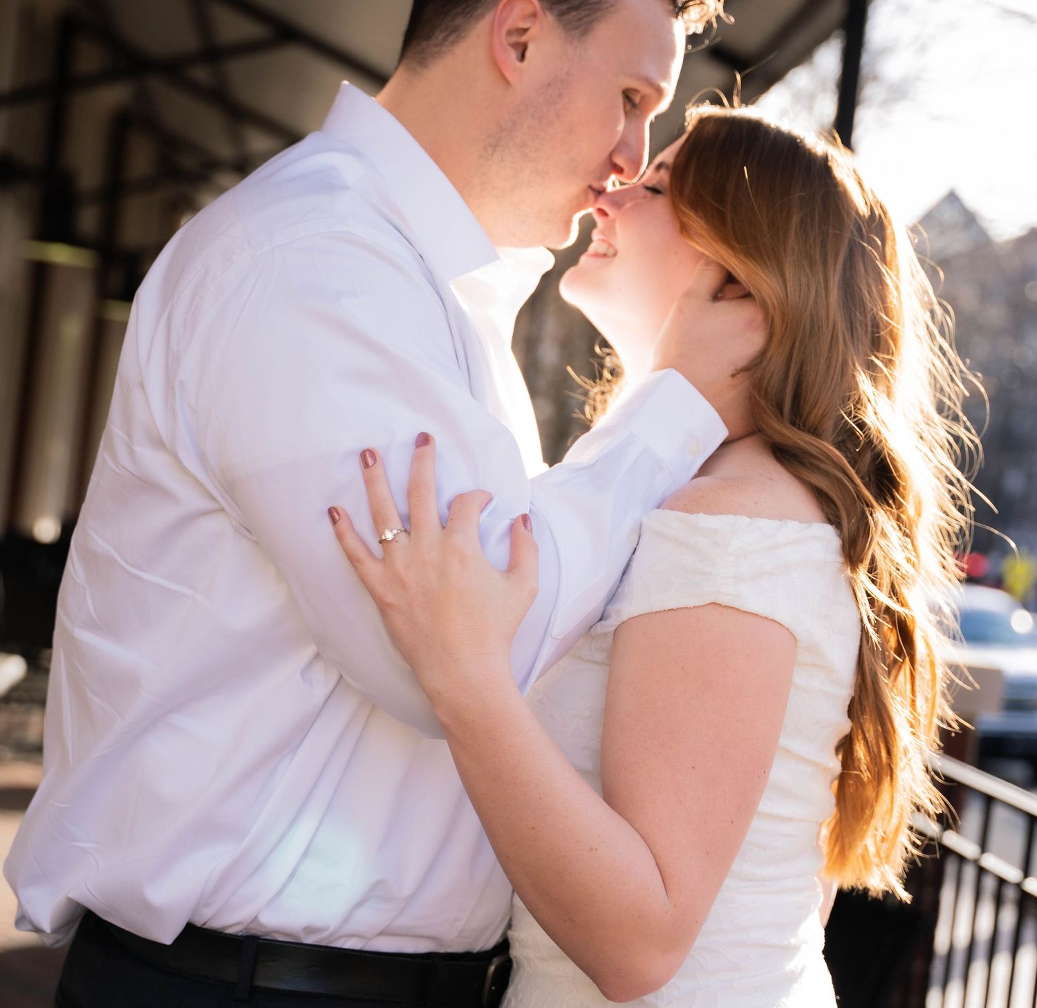 Couple sharing a romantic kiss outdoors, bride in white off-shoulder dress embracing groom in white dress shirt.