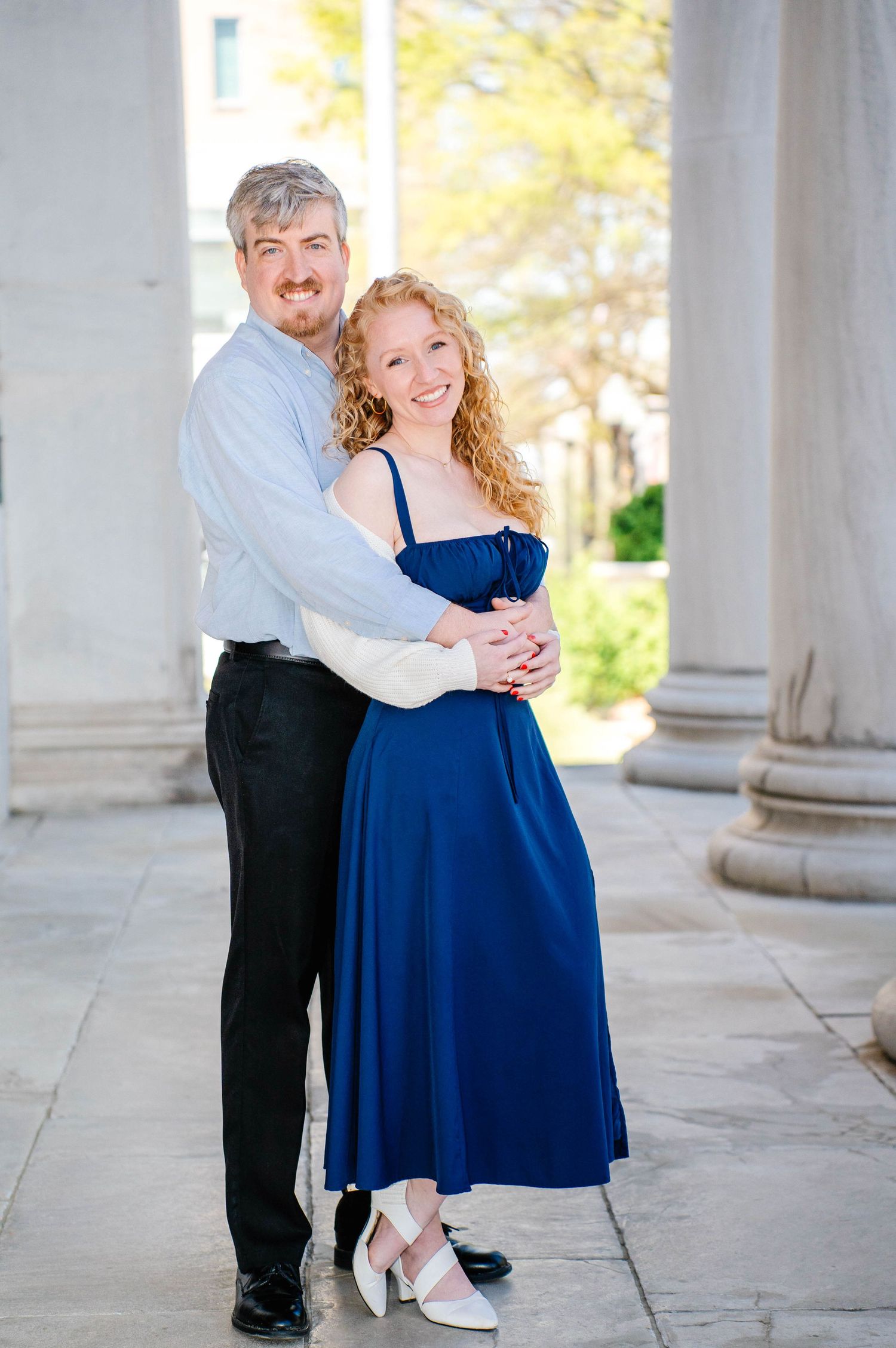A smiling couple poses together, woman in blue dress and man in light shirt embracing near elegant stone columns outdoors.