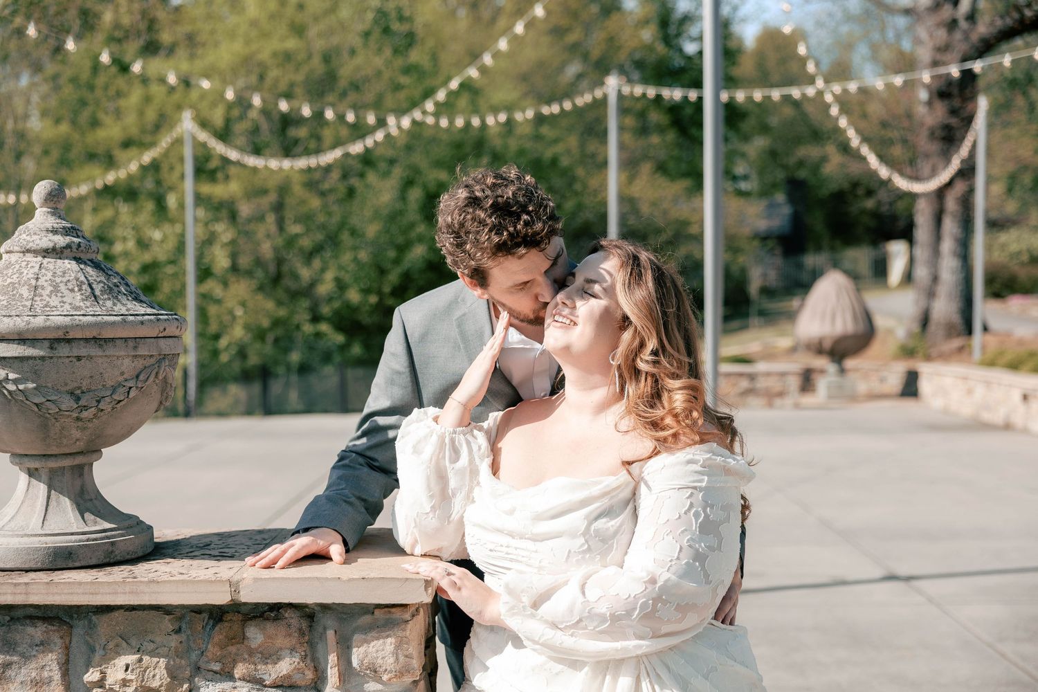 A bride and groom share a romantic moment outdoors, with string lights and stone urns in a sunlit garden setting.