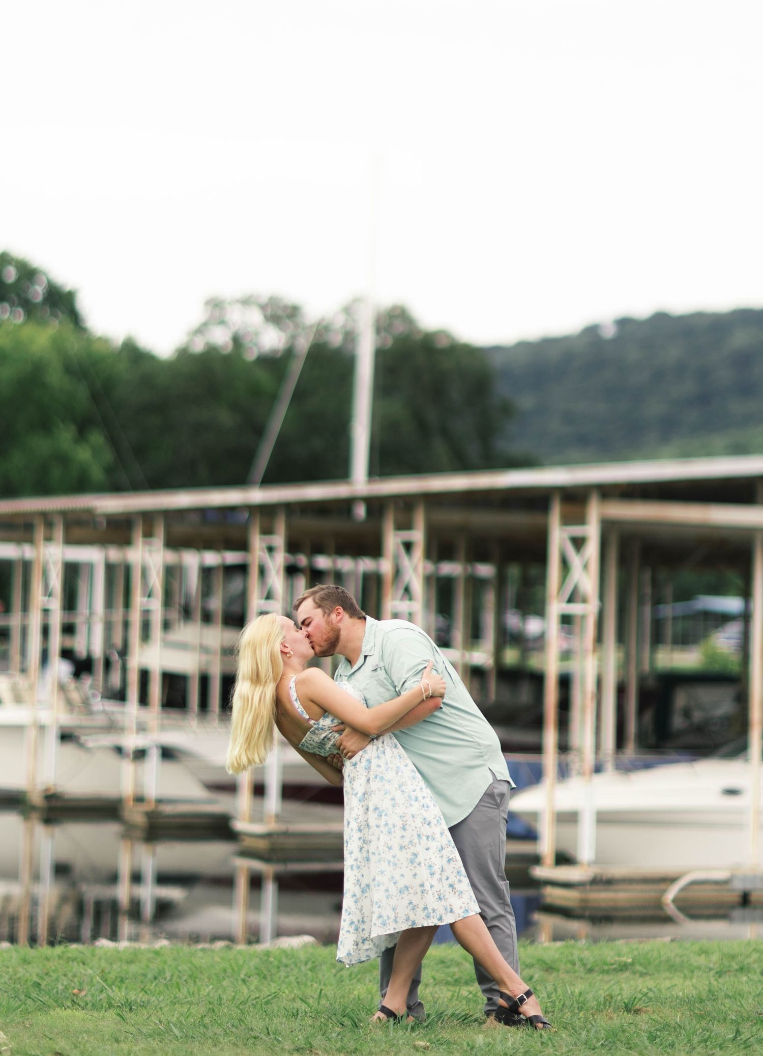 Couple sharing a romantic kiss near a marina dock with boats and green hills in the background.