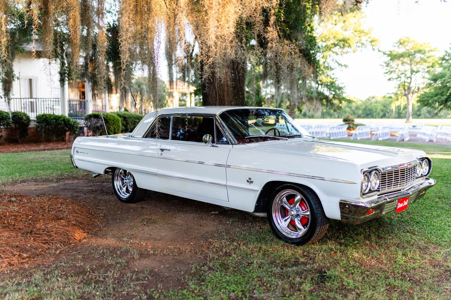 White 1964 Chevrolet Impala classic car parked on dirt path surrounded by Spanish moss-draped trees.