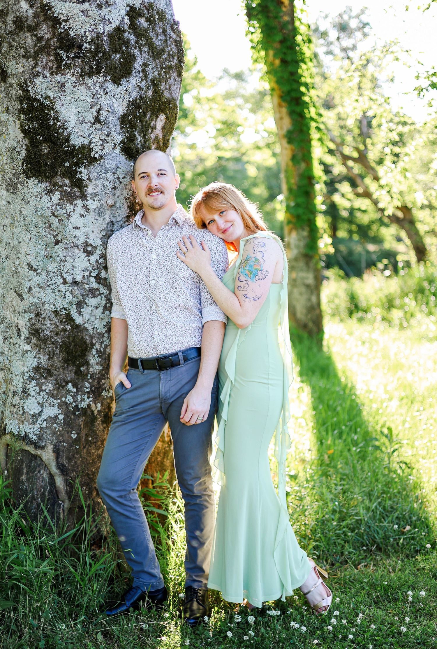 Couple posing outdoors near a mossy tree, woman in mint dress with tattoos leaning on man in floral shirt.