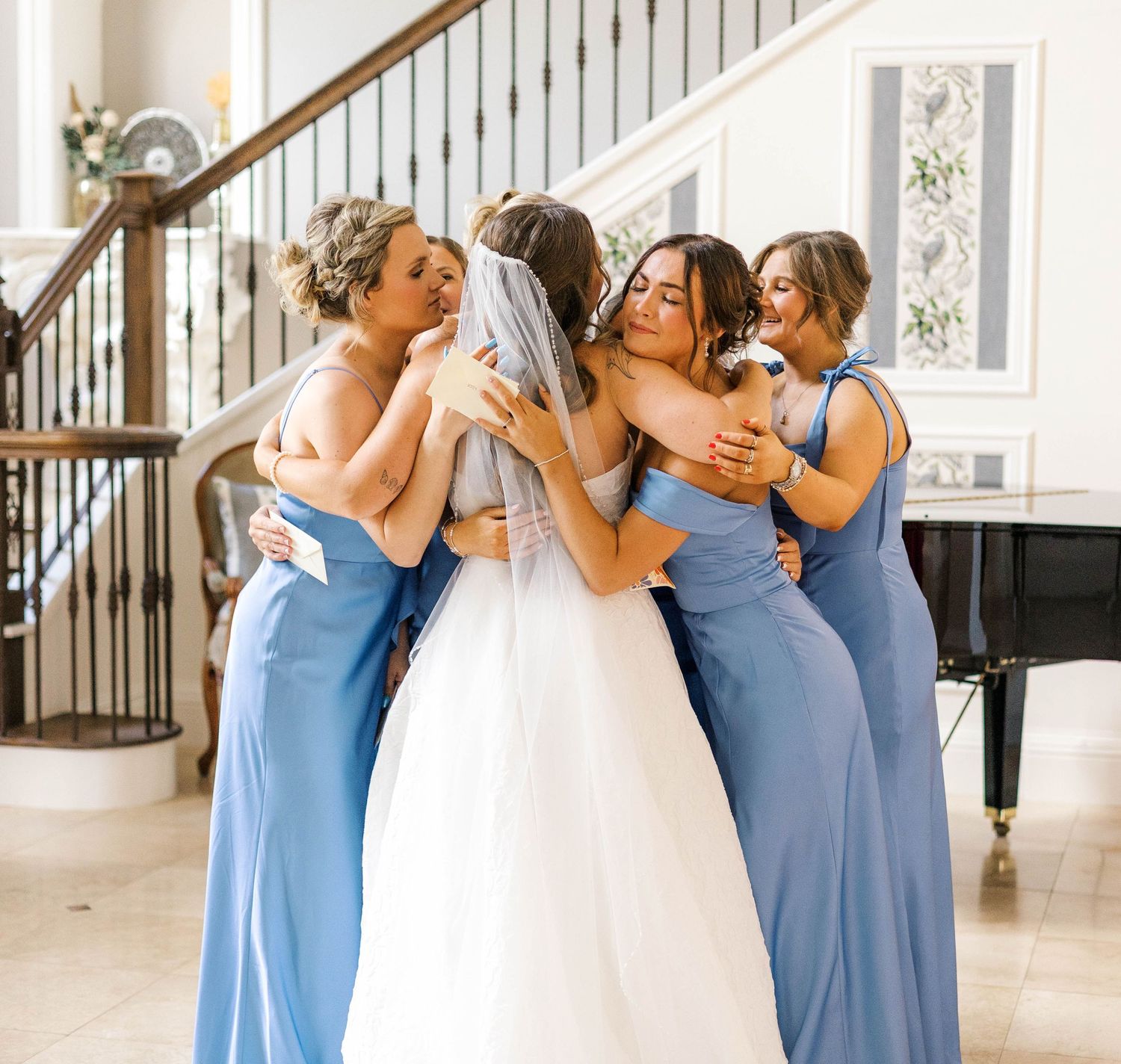 Bride in white gown hugged by bridesmaids in blue dresses near staircase indoors.