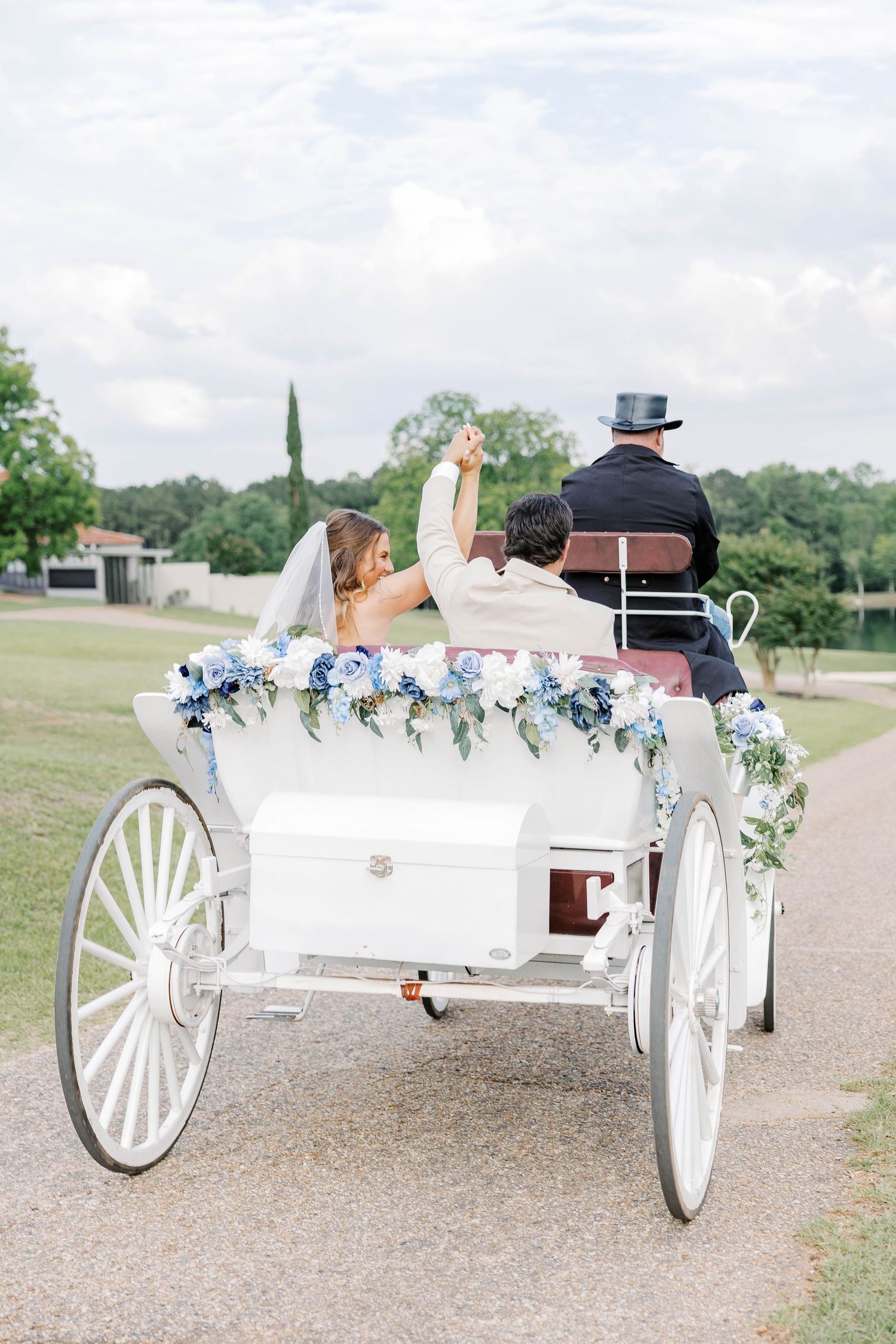 Bride and groom riding in a white horse-drawn carriage decorated with blue hydrangeas on a scenic outdoor path.