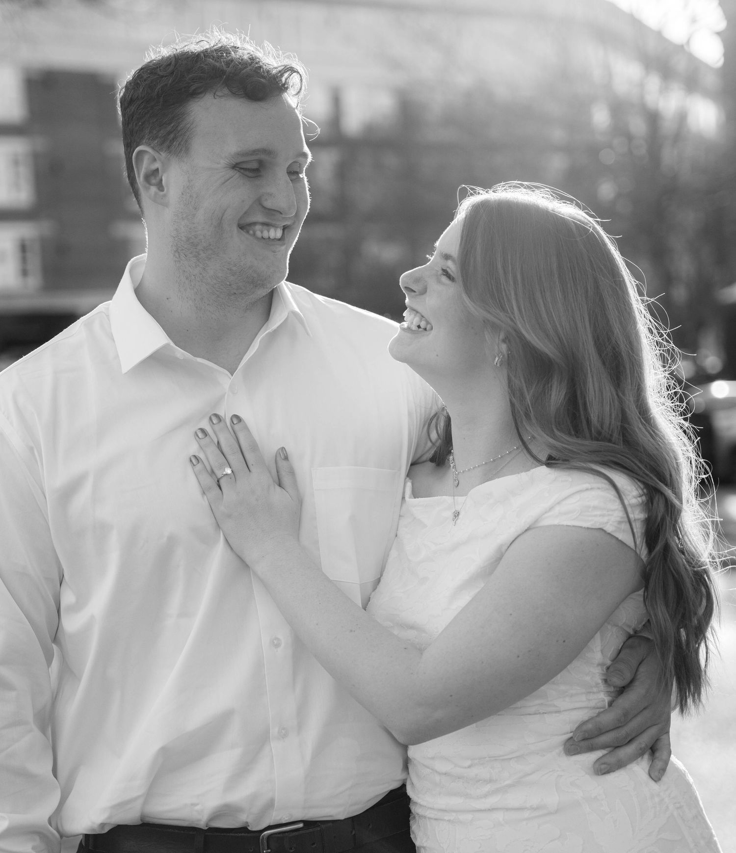 Black and white photo of a smiling couple embracing outdoors, woman wearing engagement ring, romantic portrait.