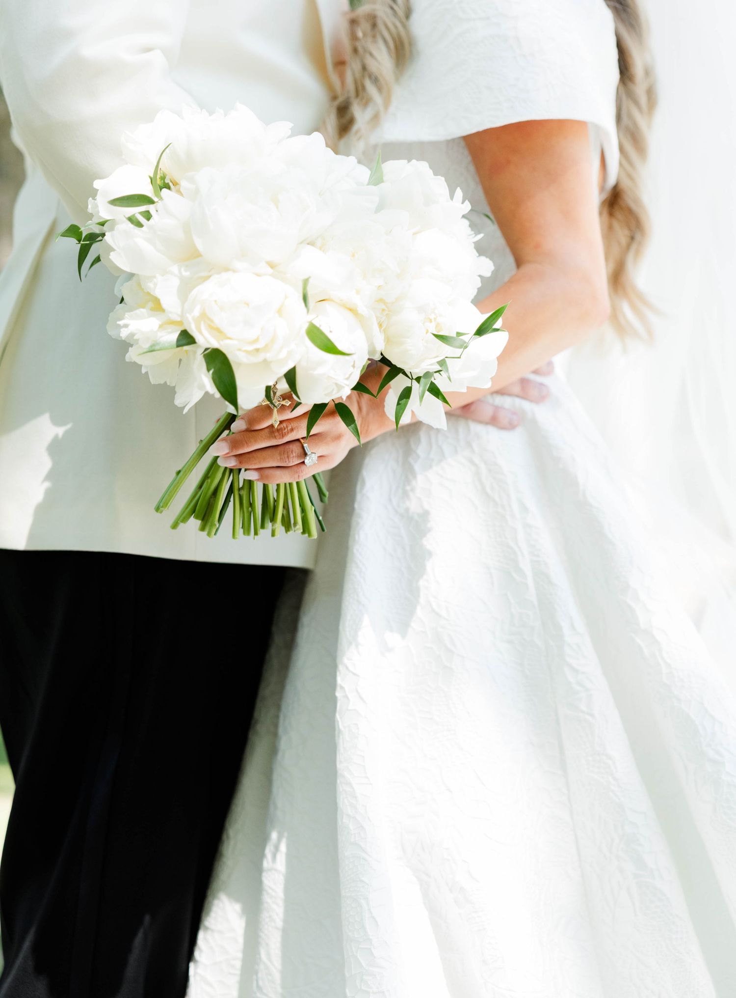 Bride holding elegant white rose and peony wedding bouquet with green stems against white bridal gown.