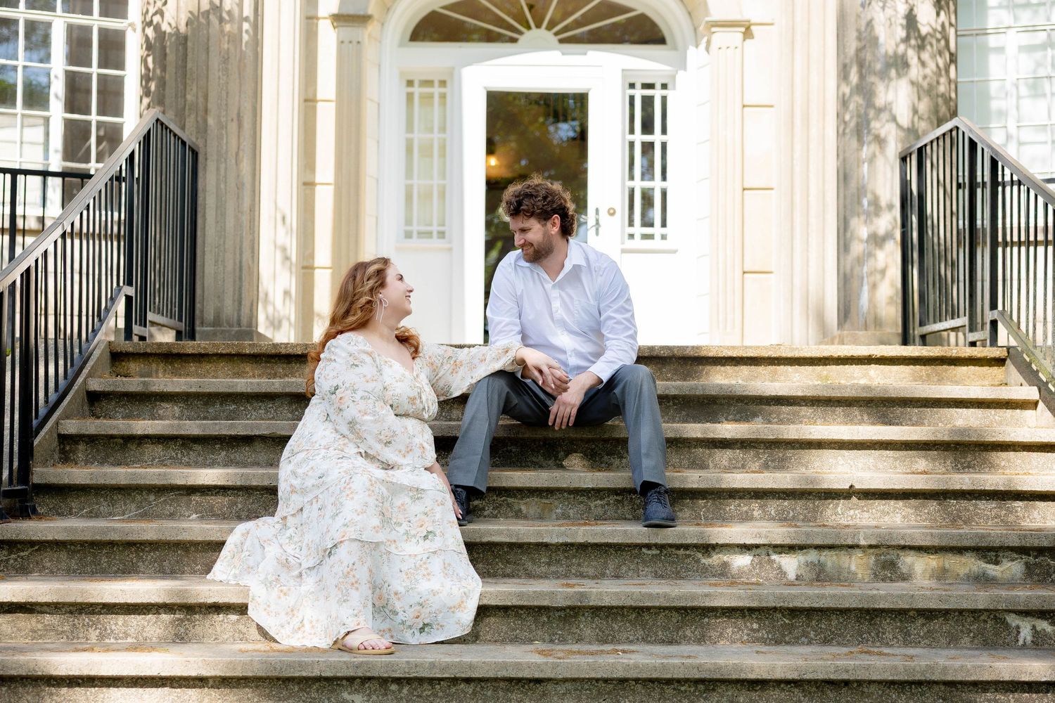 Couple sitting on stone steps of elegant building, woman in floral dress and man in casual attire, sharing a romantic moment.