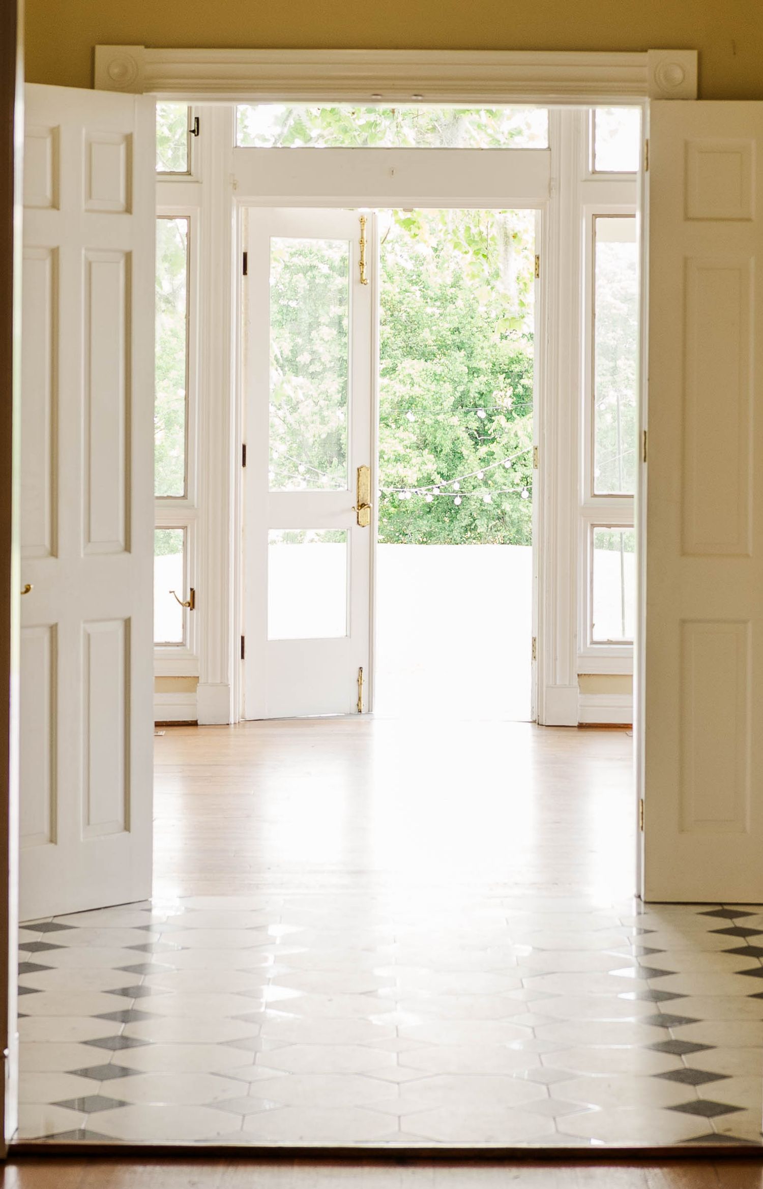 Bright white foyer with open French doors, diamond-pattern tile floor, and lush green trees visible outside.