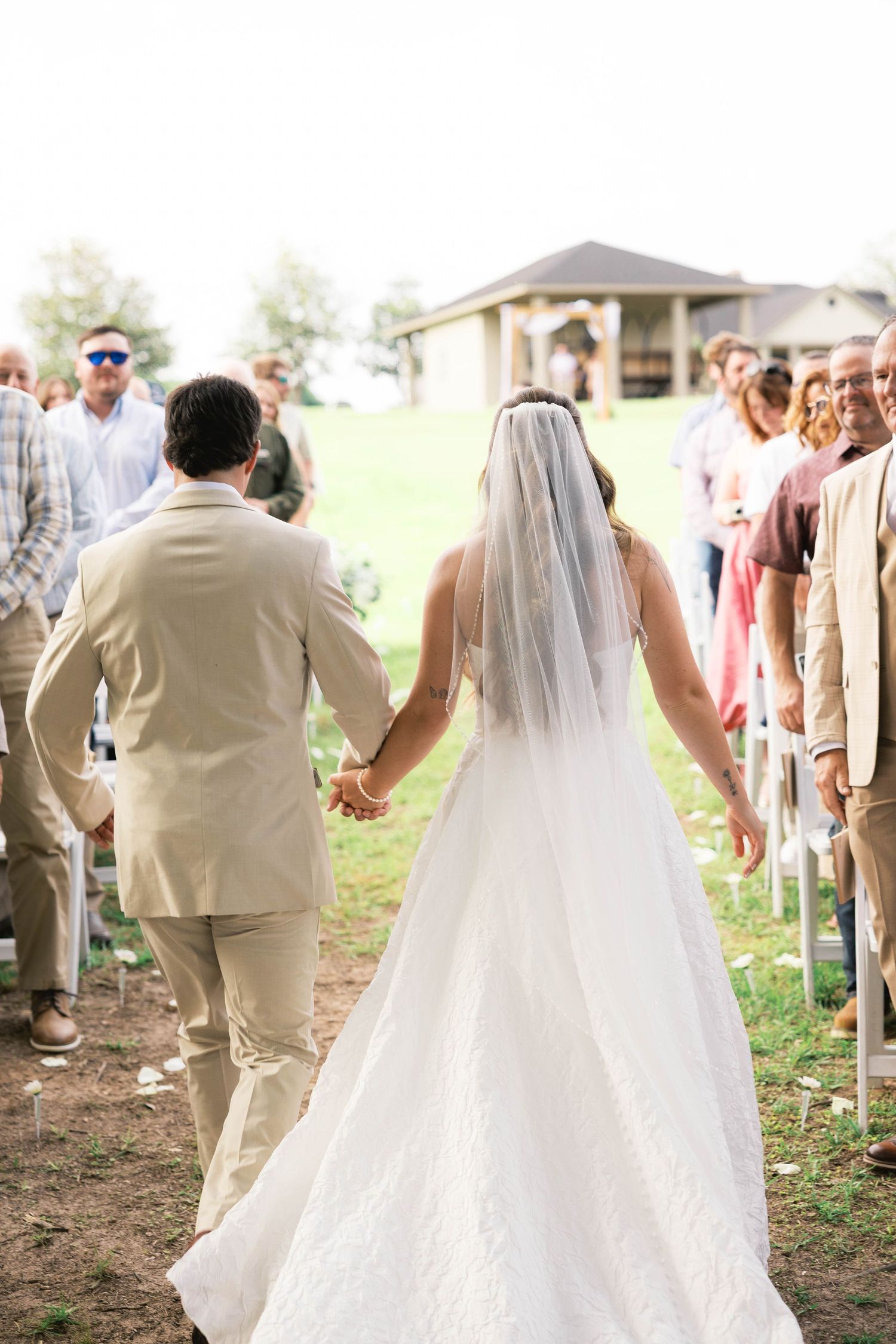 Bride and groom walk hand-in-hand down the aisle at an outdoor wedding ceremony surrounded by guests.