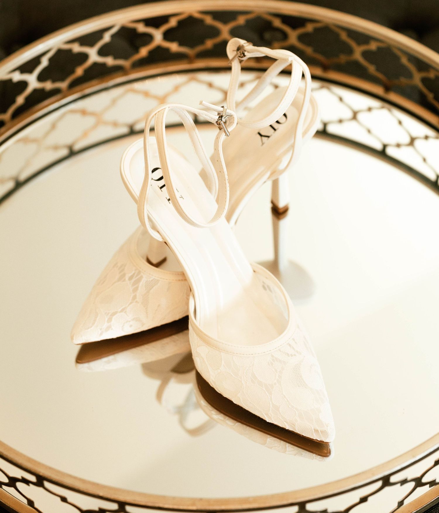 Elegant white lace bridal heels with ankle straps displayed on a mirrored tray.