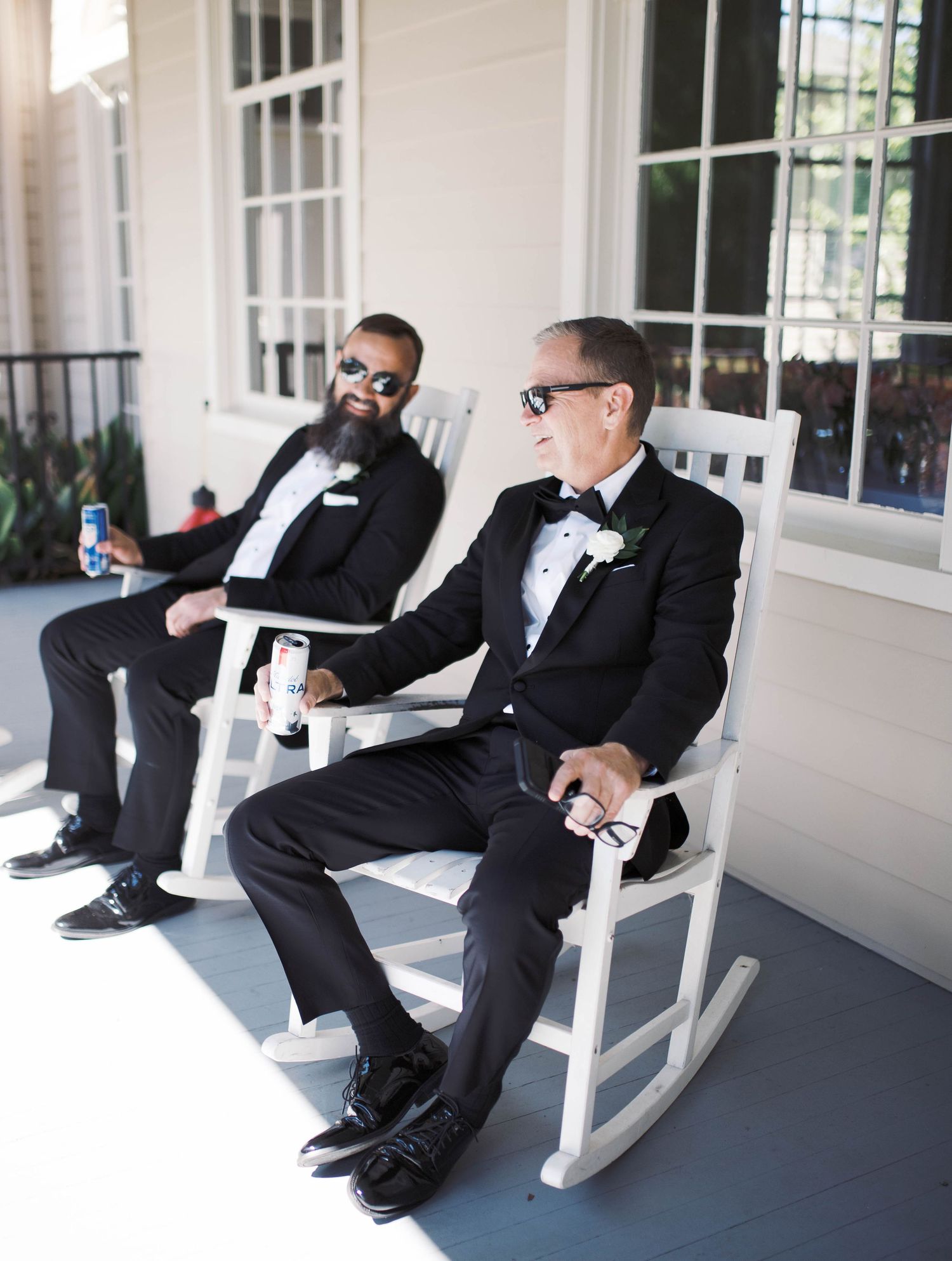 Two groomsmen in black tuxedos relax in white rocking chairs on a porch before the wedding.