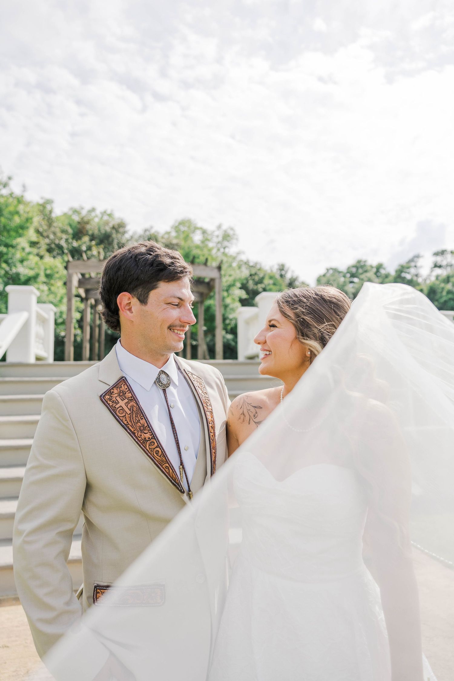 Bride and groom smiling at each other outdoors, groom in tan suit with decorative tie, bride's veil flowing in the wind.