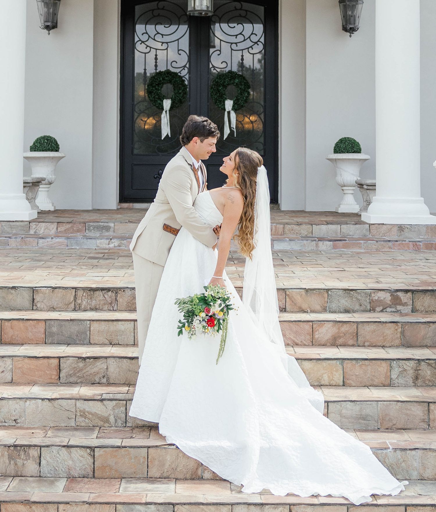 Bride and groom share a romantic moment on stone steps before elegant white mansion with wreath-adorned black doors.