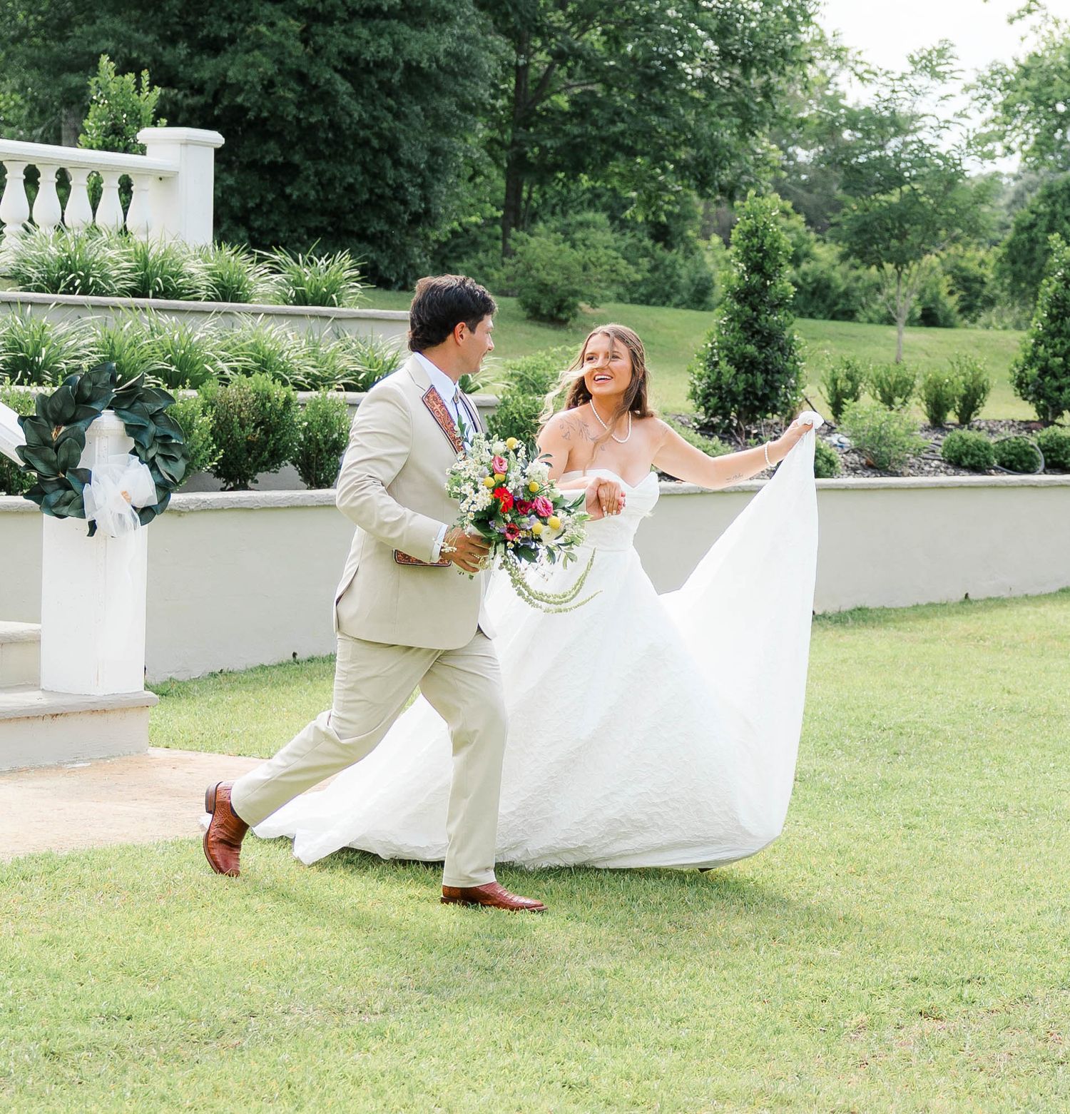 Bride in flowing white gown and groom in tan suit walk joyfully through lush garden holding colorful wedding bouquet.