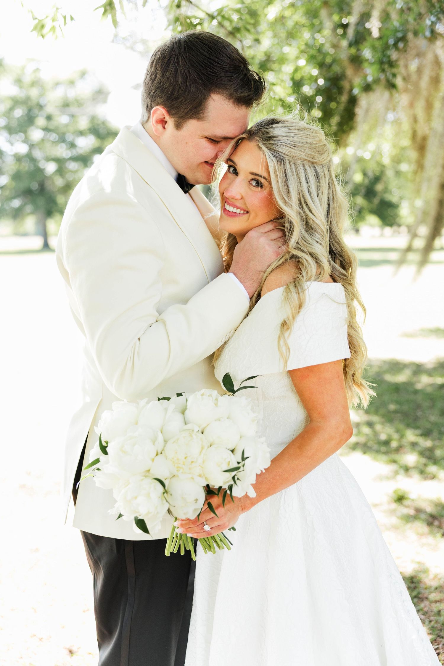 A bride and groom embrace outdoors, she holds white peony bouquet wearing wedding dress, he wears white tuxedo jacket.