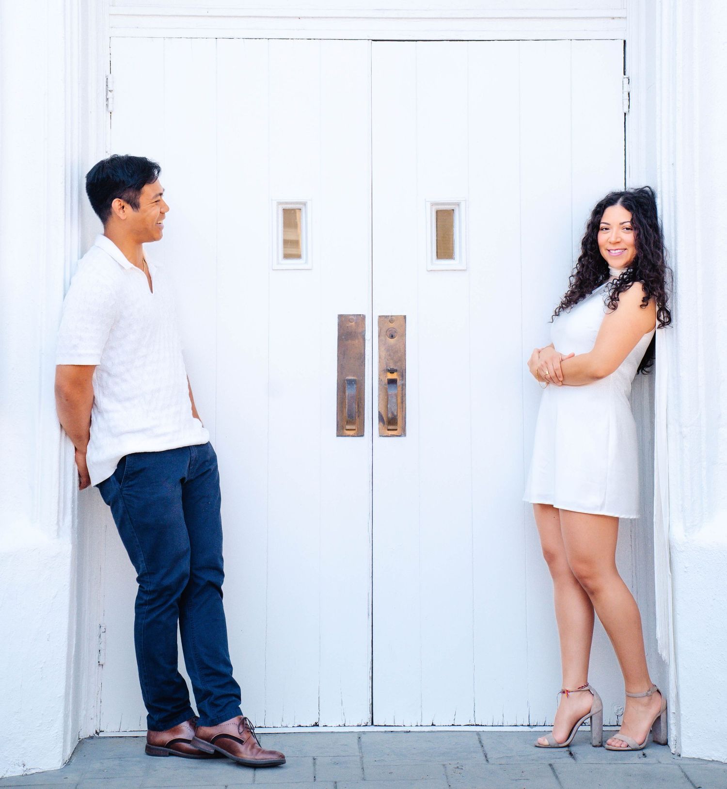 Couple in white outfits posing on either side of a white double door for an engagement photo.