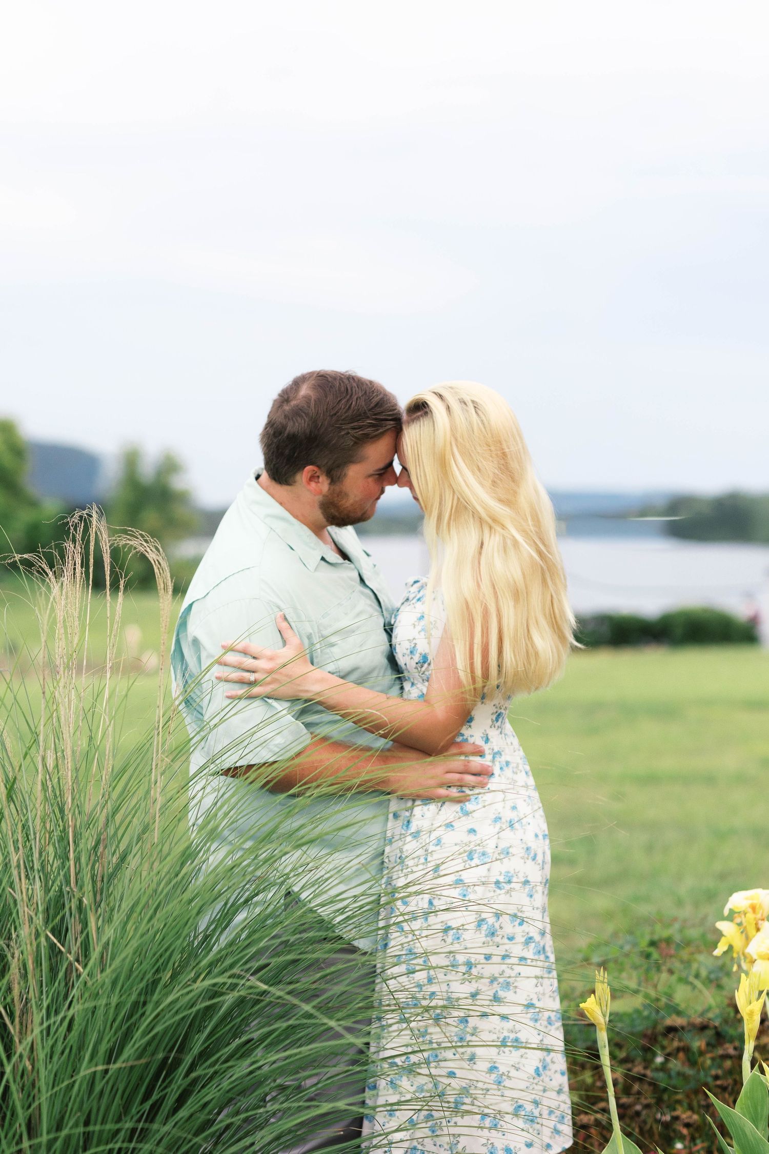 Couple embracing forehead-to-forehead in a lush green field near a lake, woman in floral dress with long blonde hair.