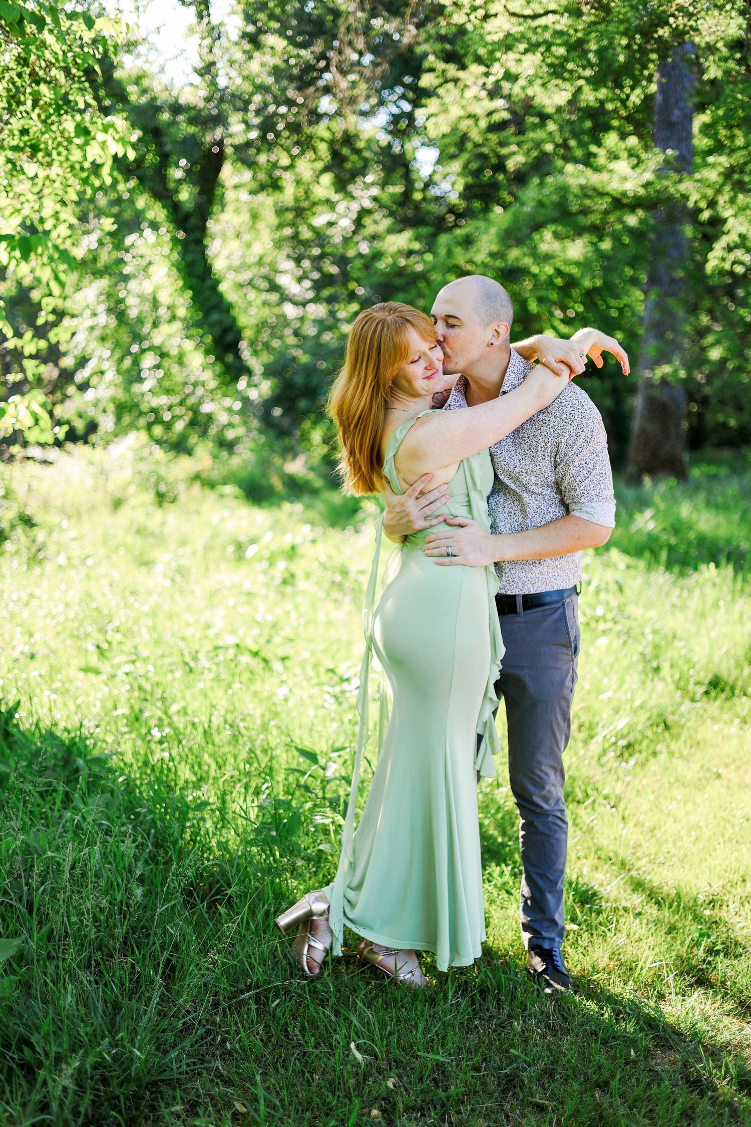 A couple embraces and kisses in a sunny green park, woman in mint dress, man in casual shirt and gray pants.