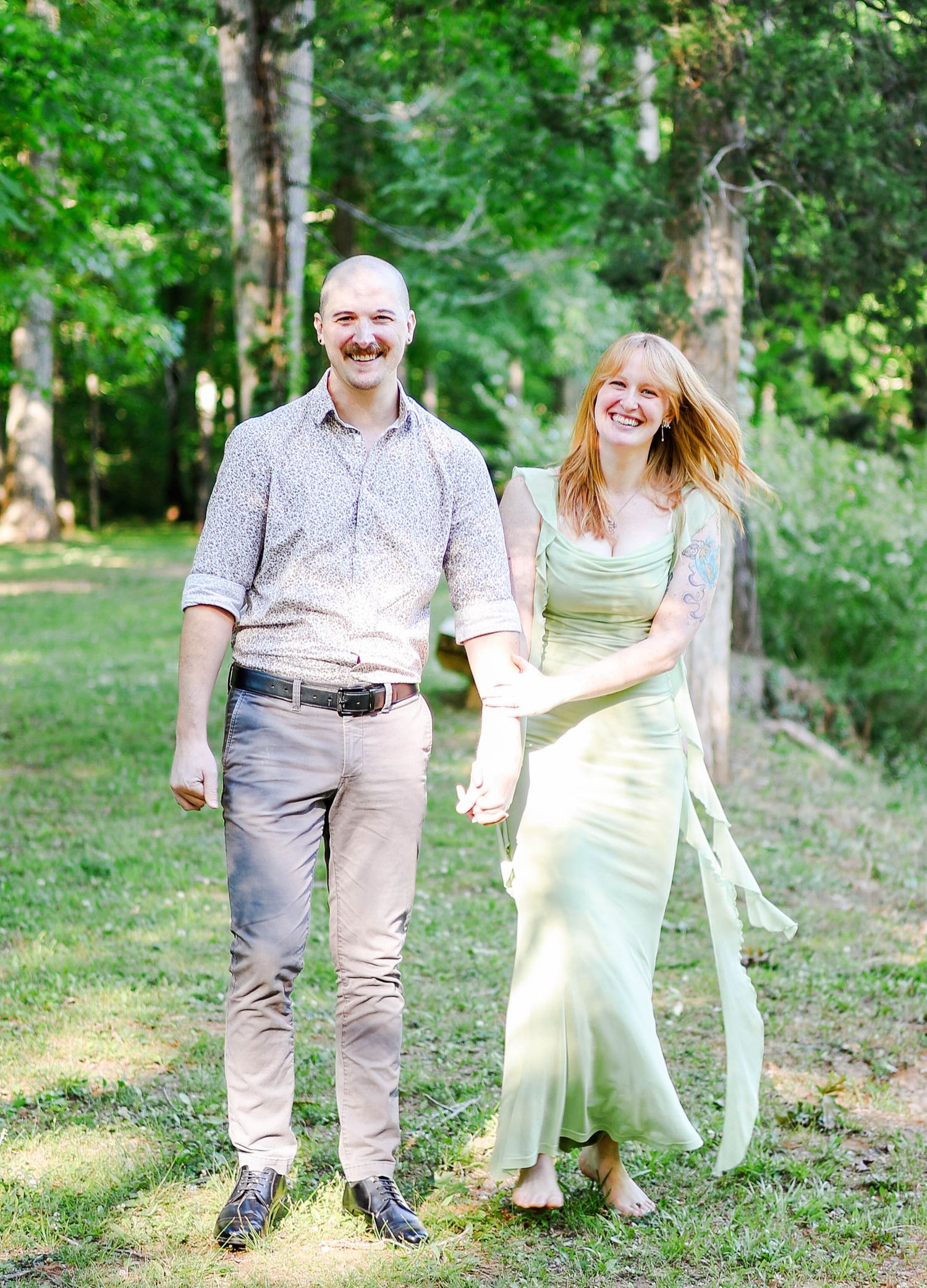 A smiling couple holds hands while walking through a lush green forest, woman in mint dress, man in casual shirt and khakis.