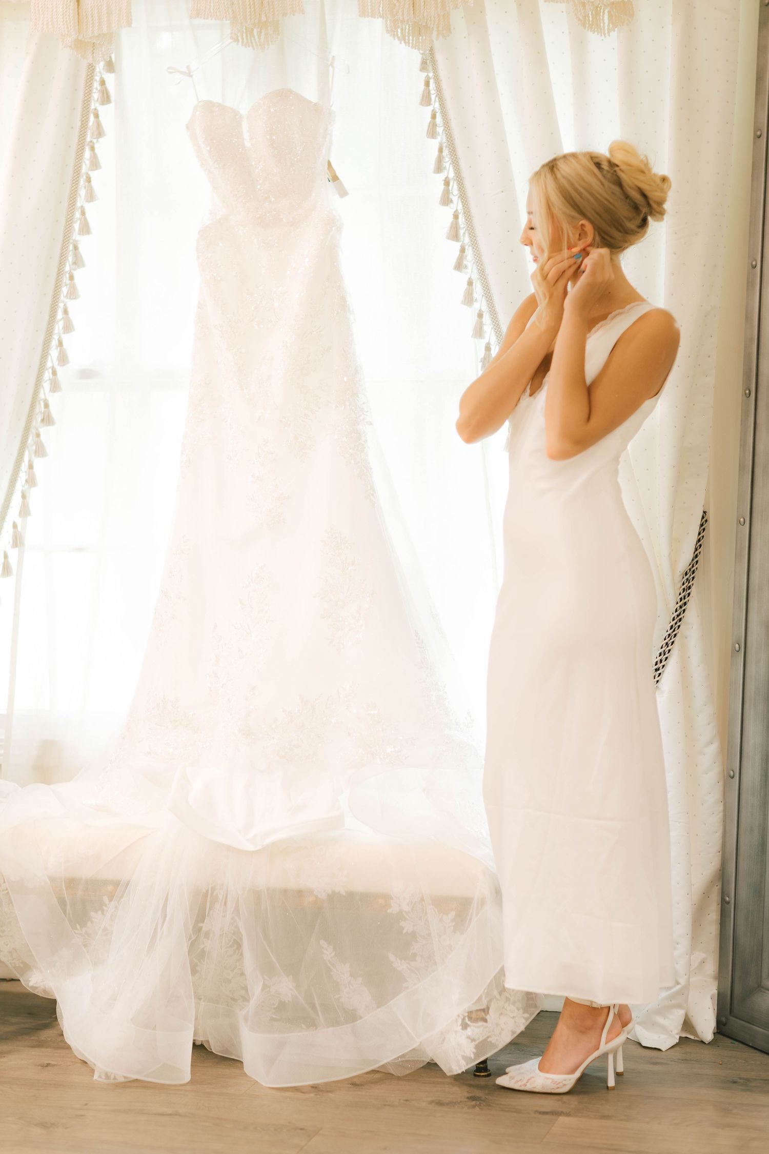 Blonde bride in white slip dress admiring her lace wedding gown hanging by a bright window.