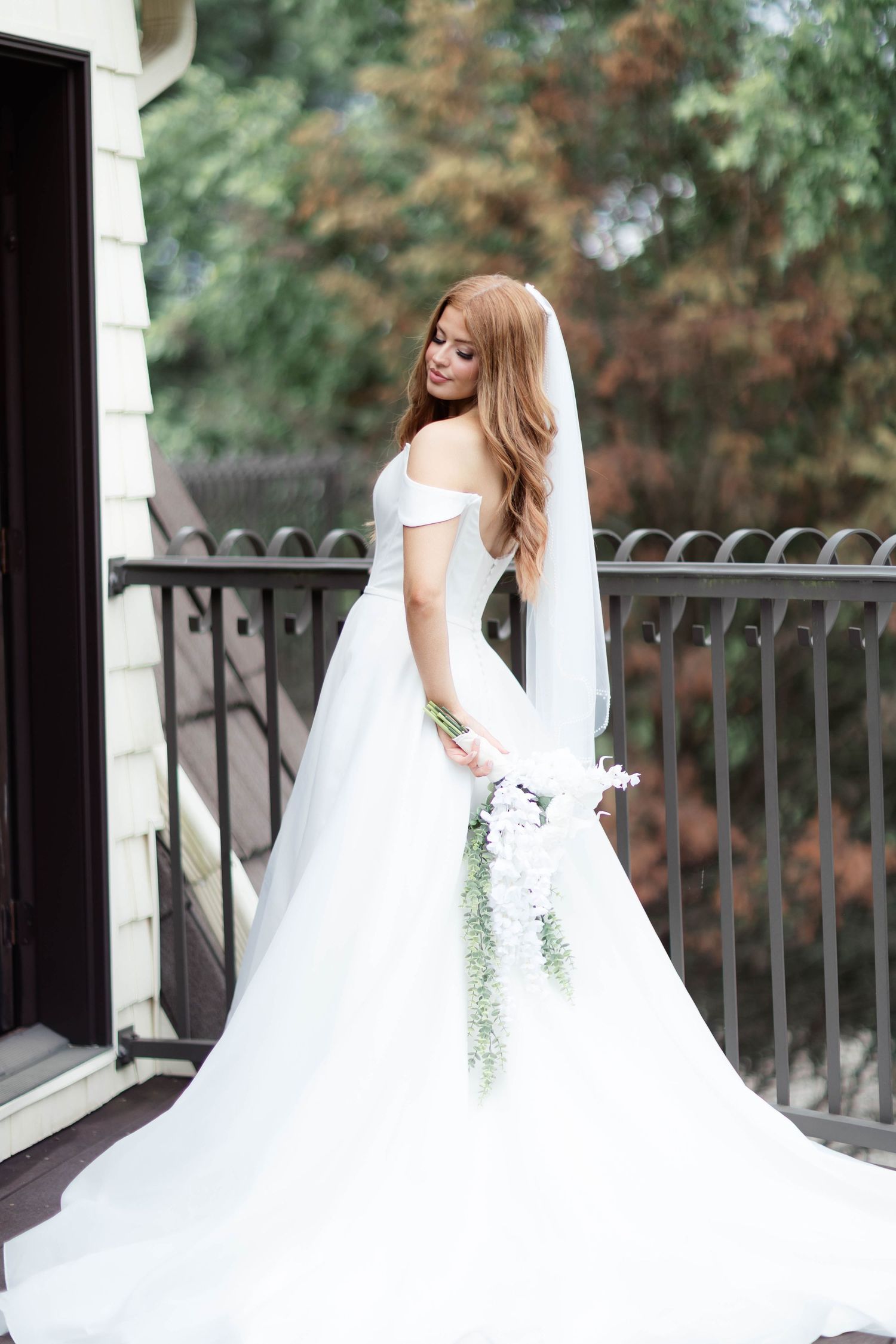 Elegant bride in white off-shoulder gown with veil holds floral bouquet on outdoor balcony.