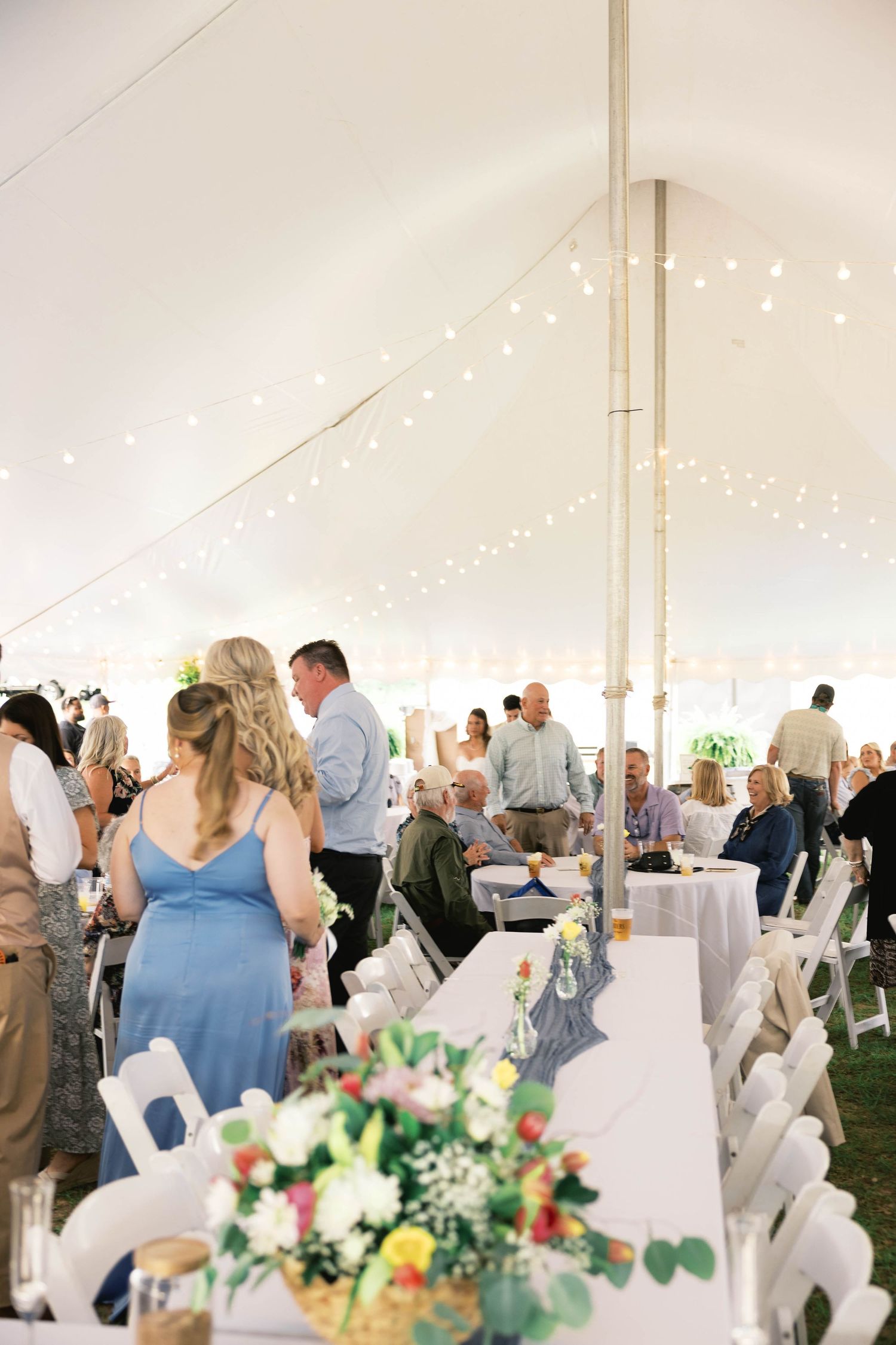 Wedding reception under white tent with string lights, guests mingling at tables with floral centerpieces.