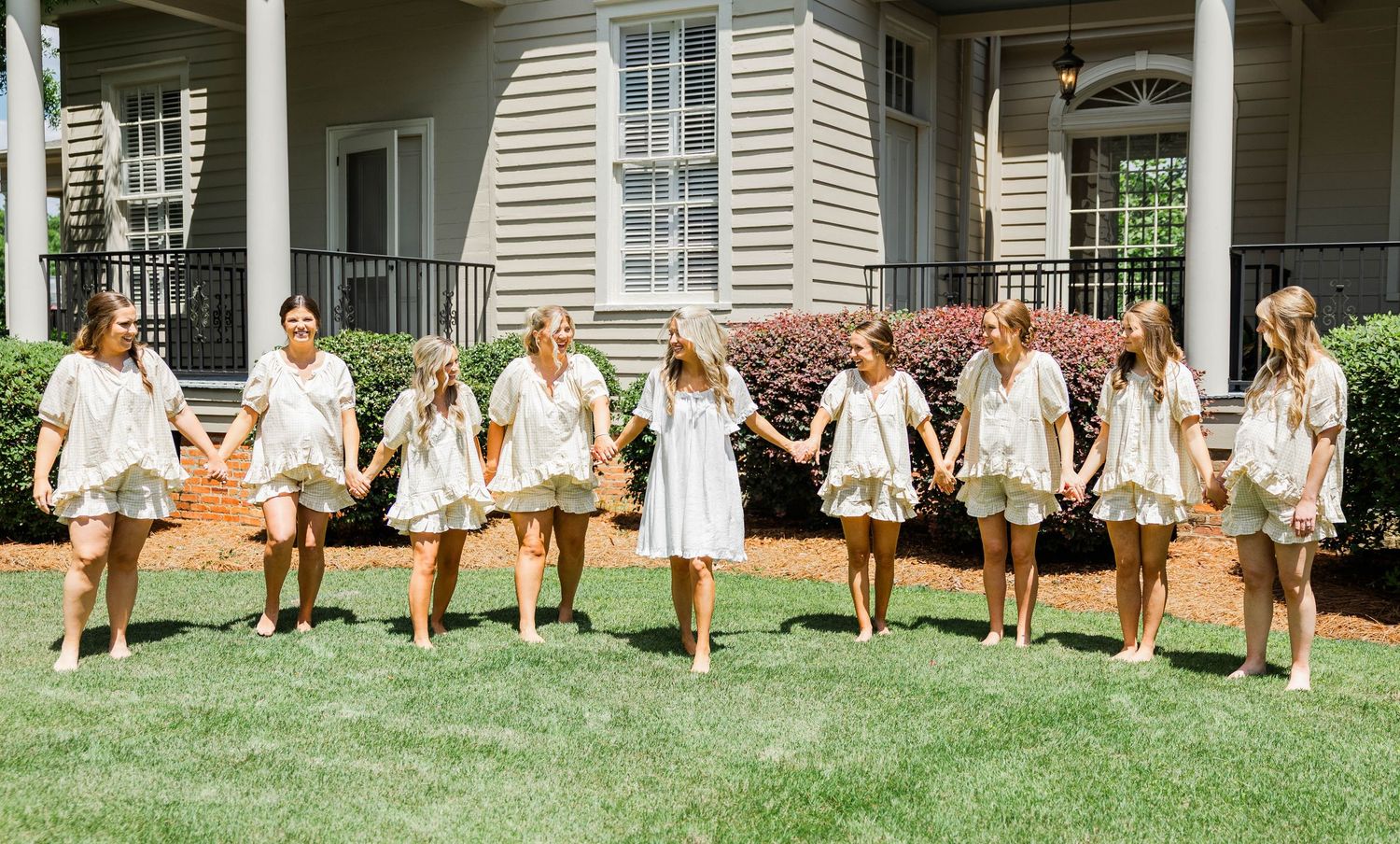 Group of girls in matching white outfits holding hands on a sunny lawn in front of a house.