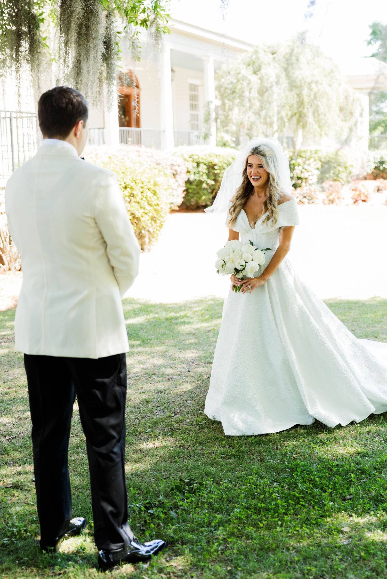 Bride in white gown with veil holds bouquet facing groom in white tuxedo jacket outdoors at elegant venue.