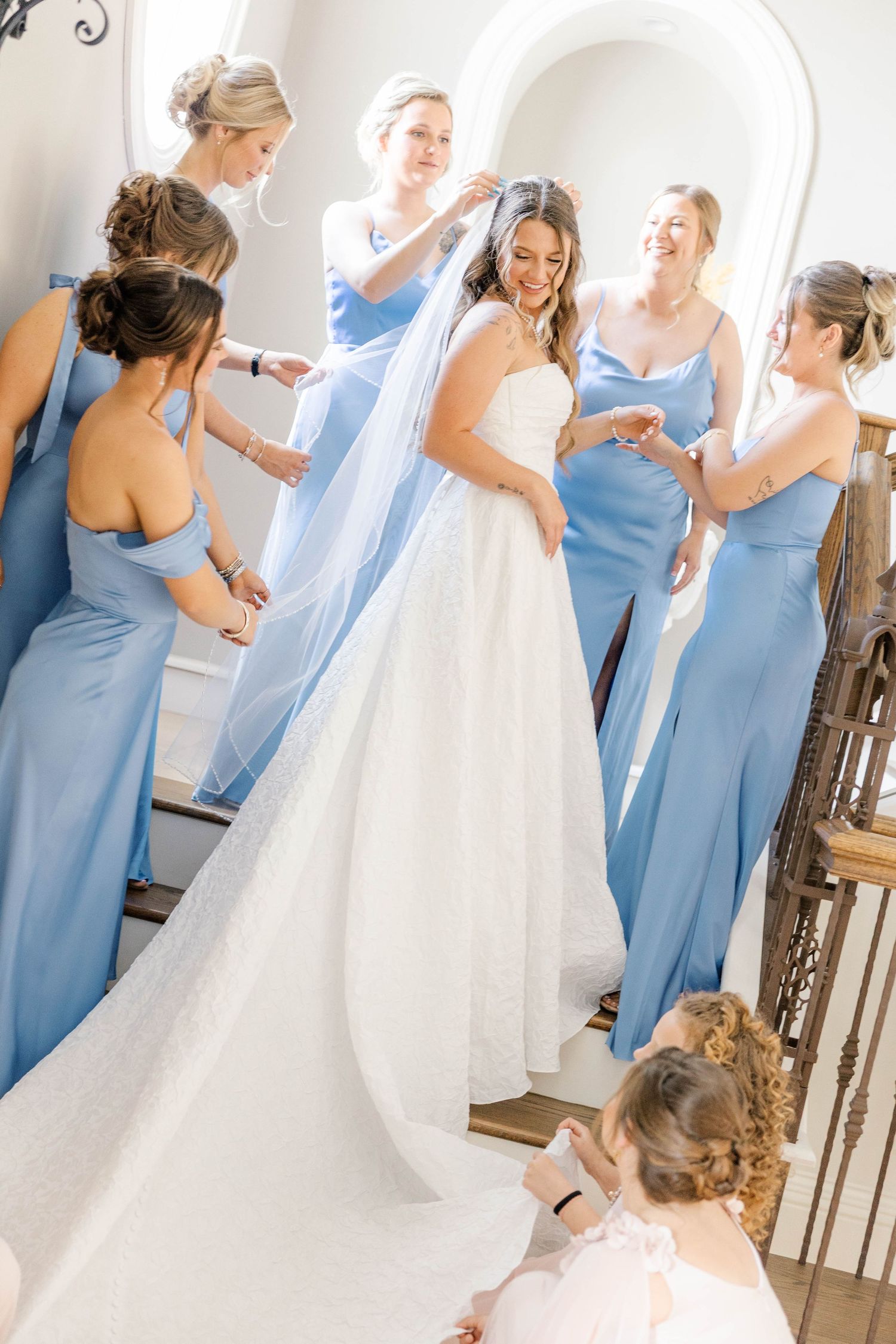 Bride in white gown surrounded by bridesmaids in blue dresses helping her prepare on a staircase.