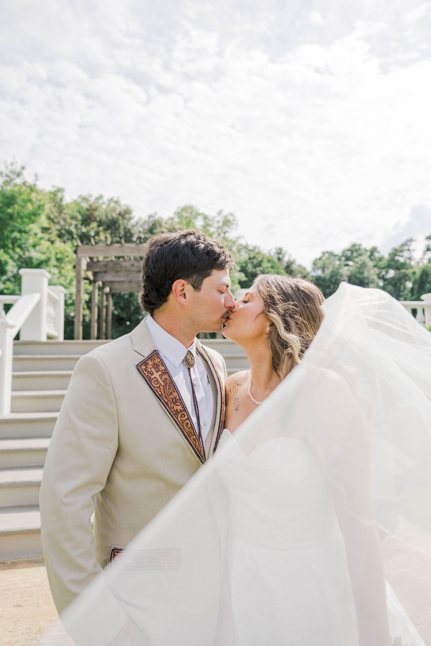 Bride and groom sharing a romantic kiss outdoors, with her white veil flowing in the breeze near a white gazebo.