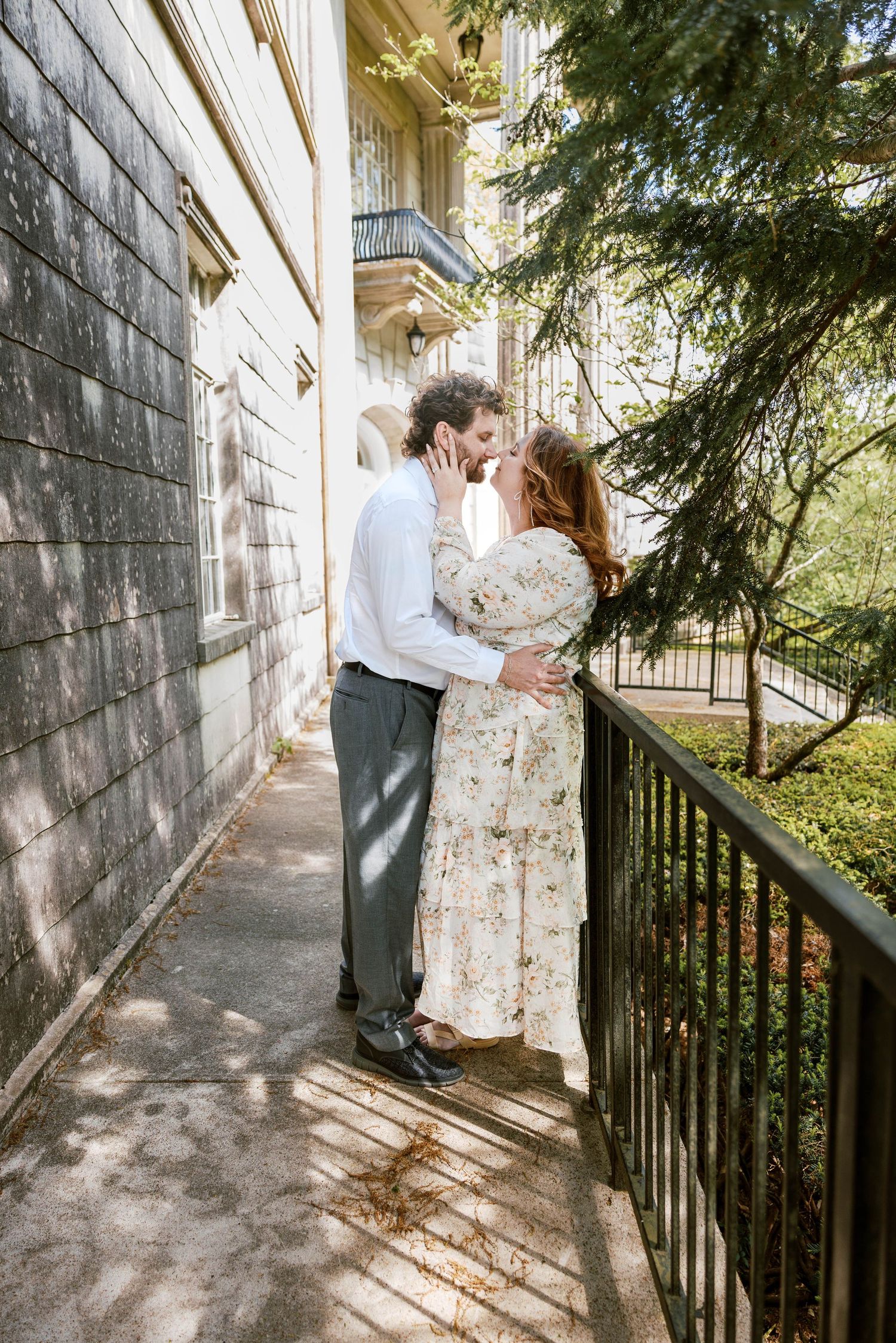 A couple shares a romantic almost-kiss on a sunlit walkway beside a building, the woman in a floral dress embracing the man.